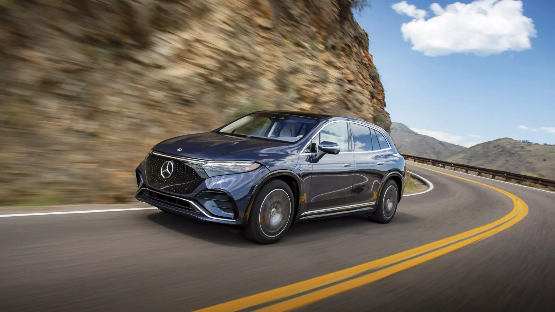 Image d'un Mercedes EQS SUV 2022 foncé en mouvement, capturée depuis une vue trois-quart avant-gauche. La voiture roule sur une route sinueuse avec une paroi rocheuse à droite. L'accent est mis sur le design luxueux avec une grille proéminente portant l'emblème de Mercedes, des phares élégants, une finition brillante et une visibilité partielle des roues en alliage. L'arrière-plan flou améliore la sensation de mouvement, soulignant les capacités de maniabilité et l'esthétique premium du véhicule dans un environnement de conduite naturel.