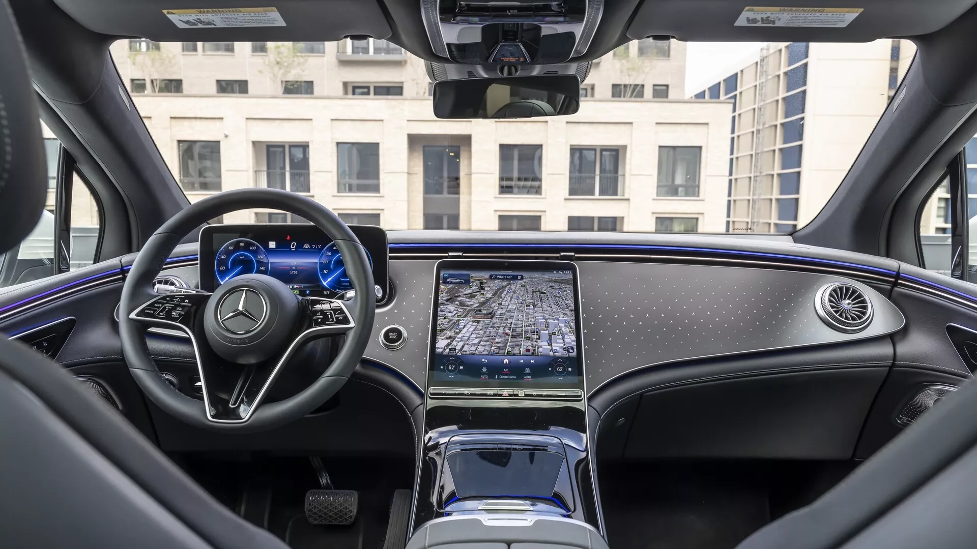 Vue intérieure du Mercedes EQS SUV 2022 depuis la perspective de la banquette arrière, montrant le tableau de bord, le volant multifonction à fond plat avec accents métalliques et la console centrale avec un système d'infodivertissement à écran tactile allongé. Le tableau de bord numérique est illuminé et le sélecteur de vitesses électronique minimaliste, les compartiments de rangement et les porte-gobelets sont visibles. L'intérieur luxueux de la voiture comprend des matériaux de haute qualité, des finitions en bois poli et un toit panoramique partiellement visible, exemplifiant le design automobile moderne et les caractéristiques technologiques avancées.