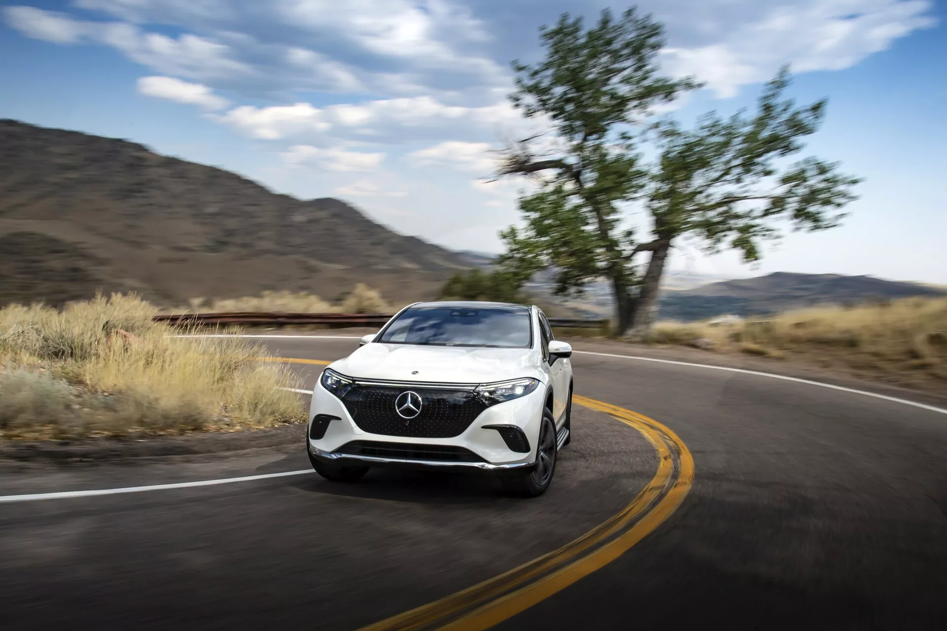 Vue avant trois quarts dynamique d'un Mercedes EQS-SUV 2022 en mouvement, présentant le design de la calandre, des phares, du capot, des roues et des lignes de contour latérales. La voiture occupe le côté droit du cadre, se déplaçant vers la gauche avec un arrière-plan flou indiquant le mouvement. Les fonctionnalités visibles incluent les feux de jour à LED, le design des phares, le pare-chocs avec prises d'air intégrées ou capteurs et les rétroviseurs latéraux aérodynamiques. La route et le paysage naturel suggèrent une utilisation sur autoroute ou en tourisme, mettant en avant les performances, le luxe et les potentielles technologies avancées du véhicule.