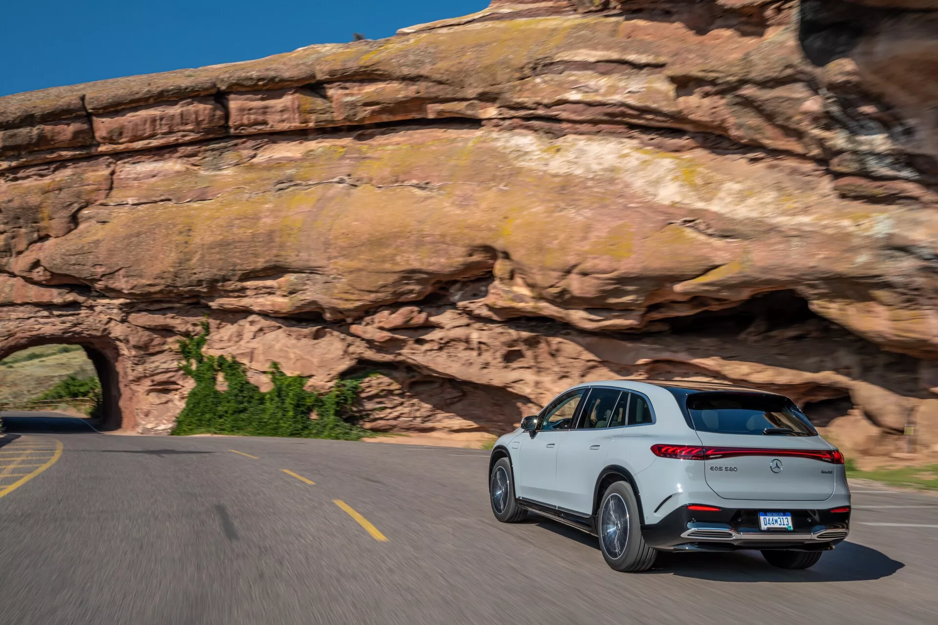 Mercedes EQS-SUV 2022 blanc roulant sur une route à deux voies sinueuse, photographié sous un angle de trois-quarts arrière, montrant ses feux arrière et le profil latéral passager. Les caractéristiques comprennent un spoiler arrière proéminent, un système d'échappement double, des garnitures argentées, des accents chromés et des roues en alliage à plusieurs branches. Le flou de mouvement sur les roues et l'arrière-plan capture la conduite dynamique du SUV dans une zone montagneuse pittoresque sous un ciel bleu clair.