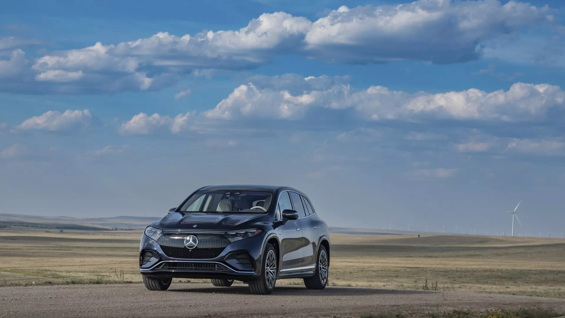 L'image montre un SUV Mercedes EQS 2022 d'un angle de trois-quarts avant, positionné sur une surface de terre plate avec un champ herbeux et des éoliennes en arrière-plan sous un ciel clair partiellement nuageux. Le véhicule, au design élégant et aérodynamique, se distingue par une calandre moderne, des phares à LED et un design de roues axé sur la performance. Les détails visibles suggèrent des systèmes avancés d'aide à la conduite, y compris des capteurs et des caméras intégrés dans le pare-chocs avant et les rétroviseurs latéraux.