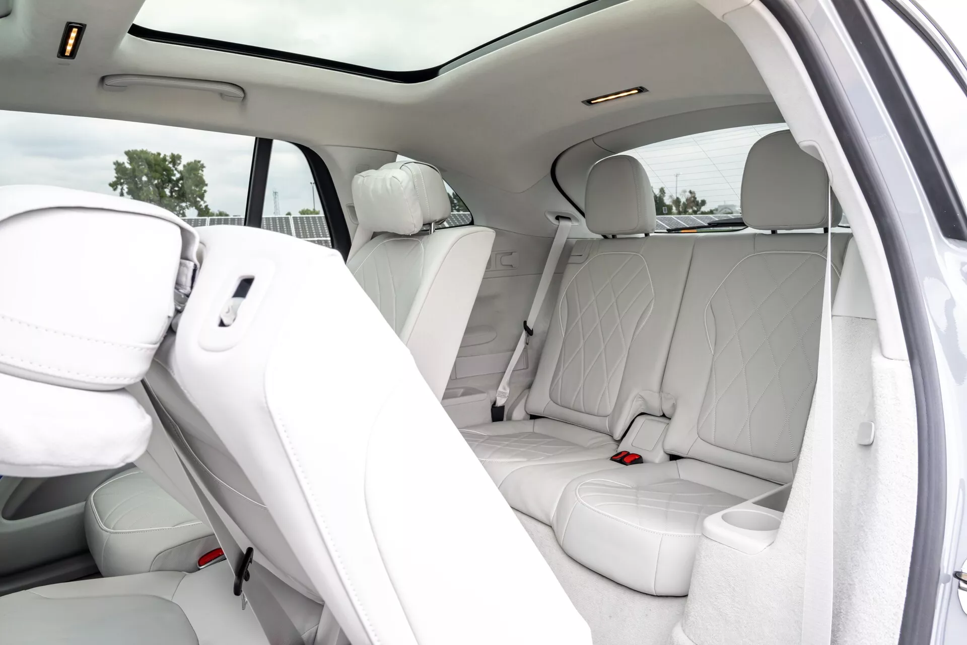Vue intérieure d'un luxueux Mercedes EQS-SUV 2022, se concentrant sur la zone des passagers arrière avec des sièges en cuir blanc perforé, un toit panoramique, un panneau de porte assorti en cuir avec des grilles de haut-parleurs, des bouches de ventilation arrière et des lumières montées au plafond. La porte ouverte révèle un aménagement de sièges spacieux et confortable pour trois passagers.