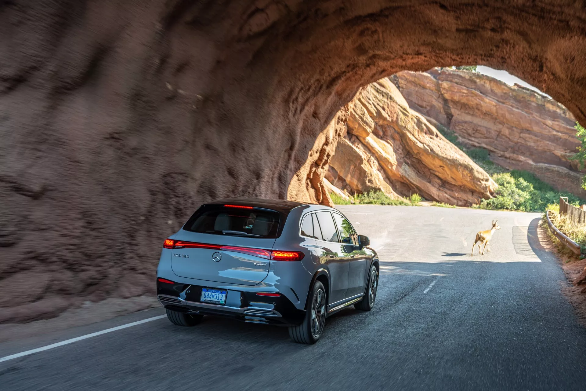 Vue arrière et angulaire d'un SUV moderne et élégant traversant un tunnel rocheux par une journée ensoleillée. Les caractéristiques clés incluent des feux arrière horizontaux enveloppants, un spoiler sportif, une antenne en aileron de requin, des garnitures de fenêtre réfléchissantes, des sorties d'échappement doubles, des roues en alliage à cinq branches et une plaque d'immatriculation montée au centre sous la vitre arrière teintée. Le véhicule montre une technologie d'éclairage avancée et inclut probablement des fonctionnalités telles qu'une caméra de recul et des capteurs de stationnement. La photo capture le mouvement et un sentiment d'exploration en plein air avec un cerf en arrière-plan.