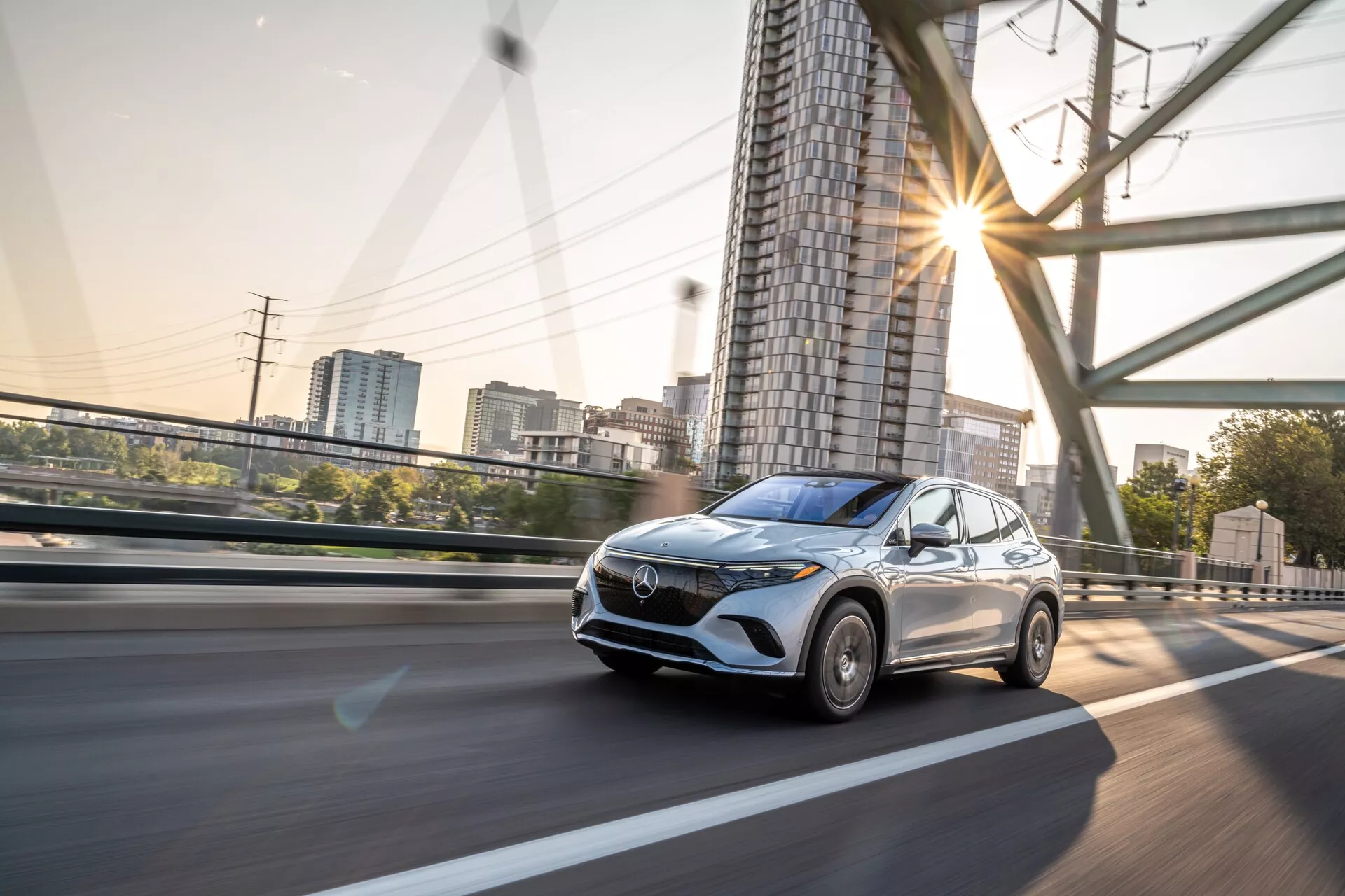 Vue dynamique d'un Mercedes EQS SUV 2022 en mouvement, capturée depuis un angle bas et une perspective avant trois quarts, montrant la calandre avant, les phares, le capot, le pare-brise et une partie du côté conducteur et du toit. La voiture roule sur une route surélevée avec des garde-corps et des structures de soutien visibles en arrière-plan. L'image met en évidence les éléments de design moderne du véhicule tels que la calandre avant proéminente, les feux de jour à LED, les lignes aérodynamiques du capot et un toit ouvrant. Le flou de mouvement sur les roues et l'arrière-plan indique une vitesse modérée à élevée, avec des reflets du soleil suggérant le matin tôt ou la fin de l'après-midi. Probablement doté de systèmes avancés d'assistance au conducteur et de régulateur de vitesse adaptatif, ce véhicule est adapté aux catégories comme les véhicules modernes, la technologie automobile, les crossovers, la dynamique des véhicules et les voitures de luxe.