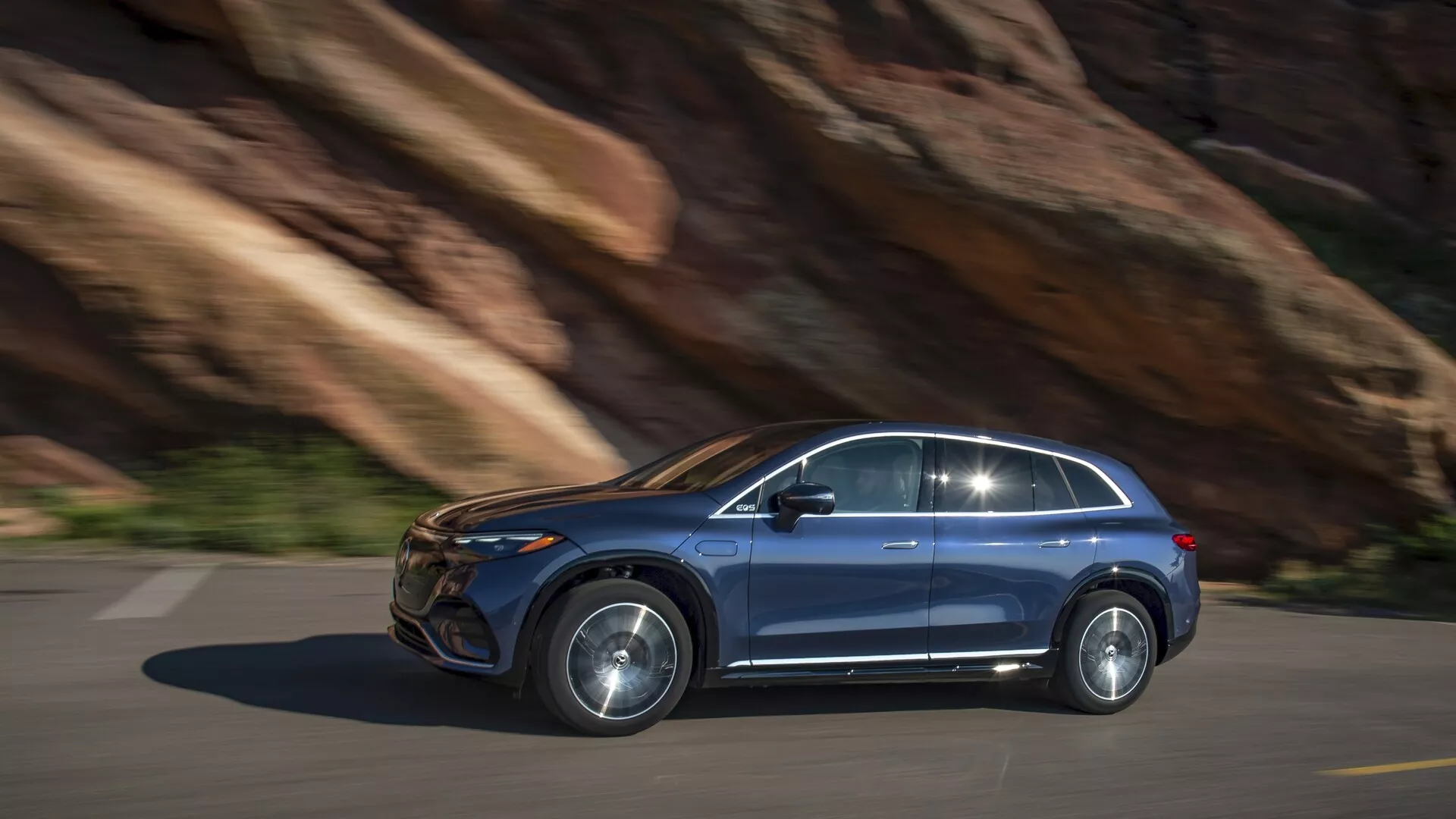 Mercedes EQS-SUV 2022 en mouvement, profil latéral. Conduisant de gauche à droite sur route pavée avec fond de roche, indiquant un terrain montagneux. Vue latérale droite montre un design moderne : portes avant et arrière visibles, fenêtres latérales bien définies avec moulure, jantes en alliage et rétroviseur latéral proéminent. Peinture métallique, probables phares LED ou HID, poignées de porte affleurantes. Possibles caractéristiques technologiques avancées : systèmes de sécurité, infotainment, moteur électrique/hybride. Légers flous de mouvement montrent une performance dynamique, photo de jour avec éclairage naturel.