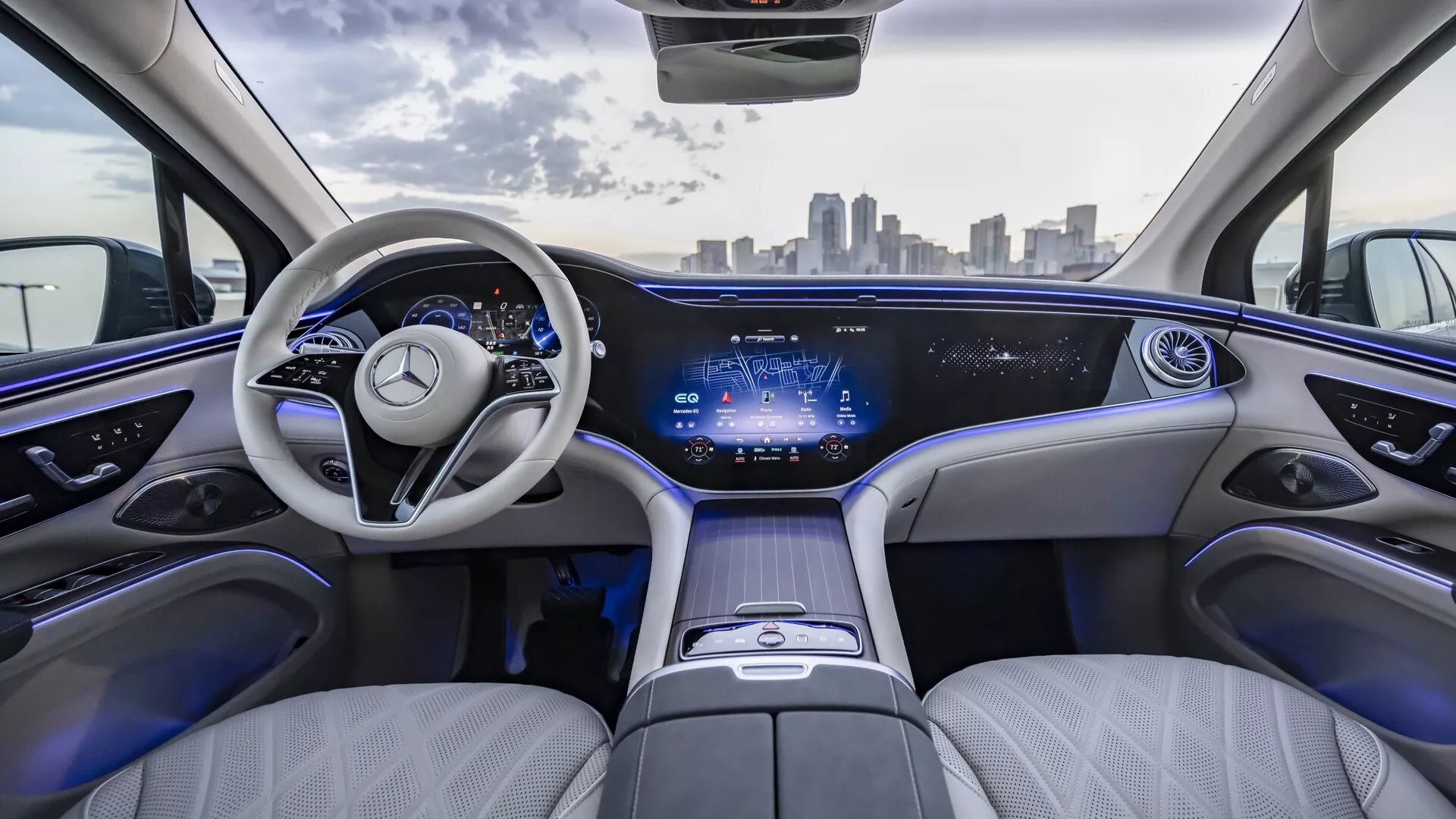 Vue intérieure du Mercedes EQS SUV 2022 depuis les sièges arrière, avec une sellerie en cuir léger de luxe, un tableau de bord élégant avec un affichage numérique continu, un volant moderne à deux branches avec l'emblème Mercedes-Benz, une disposition des sièges spacieuse et confortable, un éclairage d'ambiance, une console centrale avec commandes tactiles et un toit ouvrant panoramique. Skyline urbaine visible à travers le pare-brise.