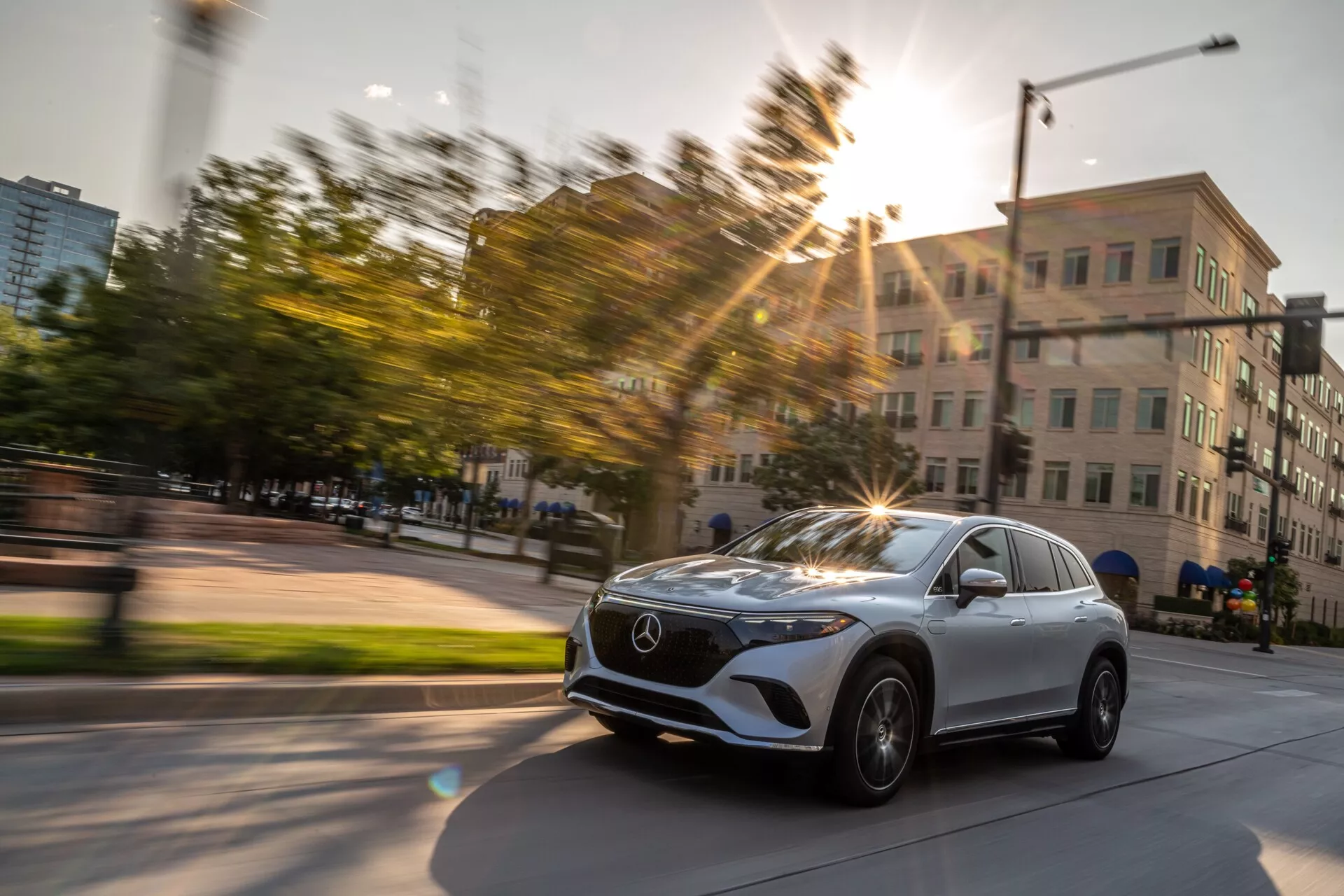 Image dynamique du Mercedes EQS SUV 2022 capturée en mouvement avec une vue de trois quarts avant légèrement diagonale. La photographie met en avant la calandre avant du véhicule avec l'emblème Mercedes, les phares LED élégants, la roue et l'aile du côté conducteur, ainsi qu'une partie du pare-brise et de la porte du conducteur. Les roues de la voiture sont tournées vers la droite, suggérant un virage ou un changement de direction. La lumière du soleil crée un effet de flare, ajoutant de l'énergie à la photo, prise en milieu urbain avec des bâtiments et de la verdure, pendant l'heure dorée. Le flou de mouvement en arrière-plan souligne la vitesse et le mouvement.