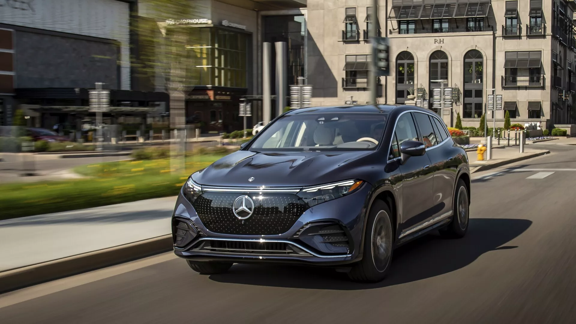 Image dynamique au niveau de la rue du Mercedes EQS SUV 2022 en mouvement, capturée sous un angle de trois quarts avant bas sur le côté conducteur. Le véhicule luxueux met en avant sa calandre frontale proéminente avec un grand emblème, des phares LED élégants avec feux de jour, et un design aérodynamique. Les roues alliage et les rétroviseurs latéraux de la couleur de la carrosserie avec clignotants intégrés soulignent le luxe. Le fond urbain avec flou de mouvement met en valeur les caractéristiques technologiques avancées de la voiture et sa présence sur la route.