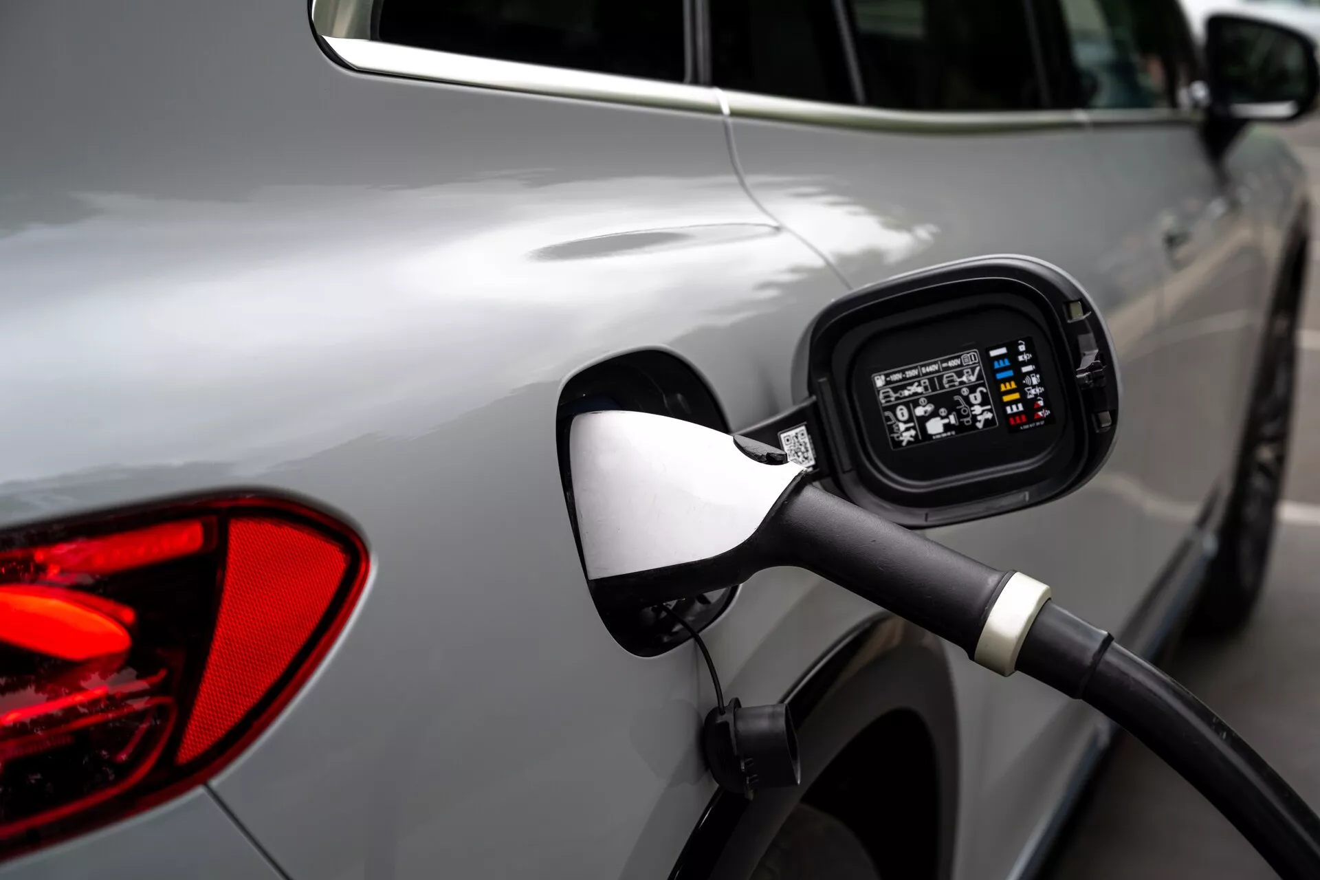 Gros plan de la zone du port de charge sur le côté d'un Mercedes EQS-SUV argenté de 2022, se concentrant sur le câble de charge branché, le port ouvert révélant un écran numérique indiquant l'état de charge, avec un arrière-plan légèrement flou montrant des parties du feu arrière et le reflet d'un autre véhicule.