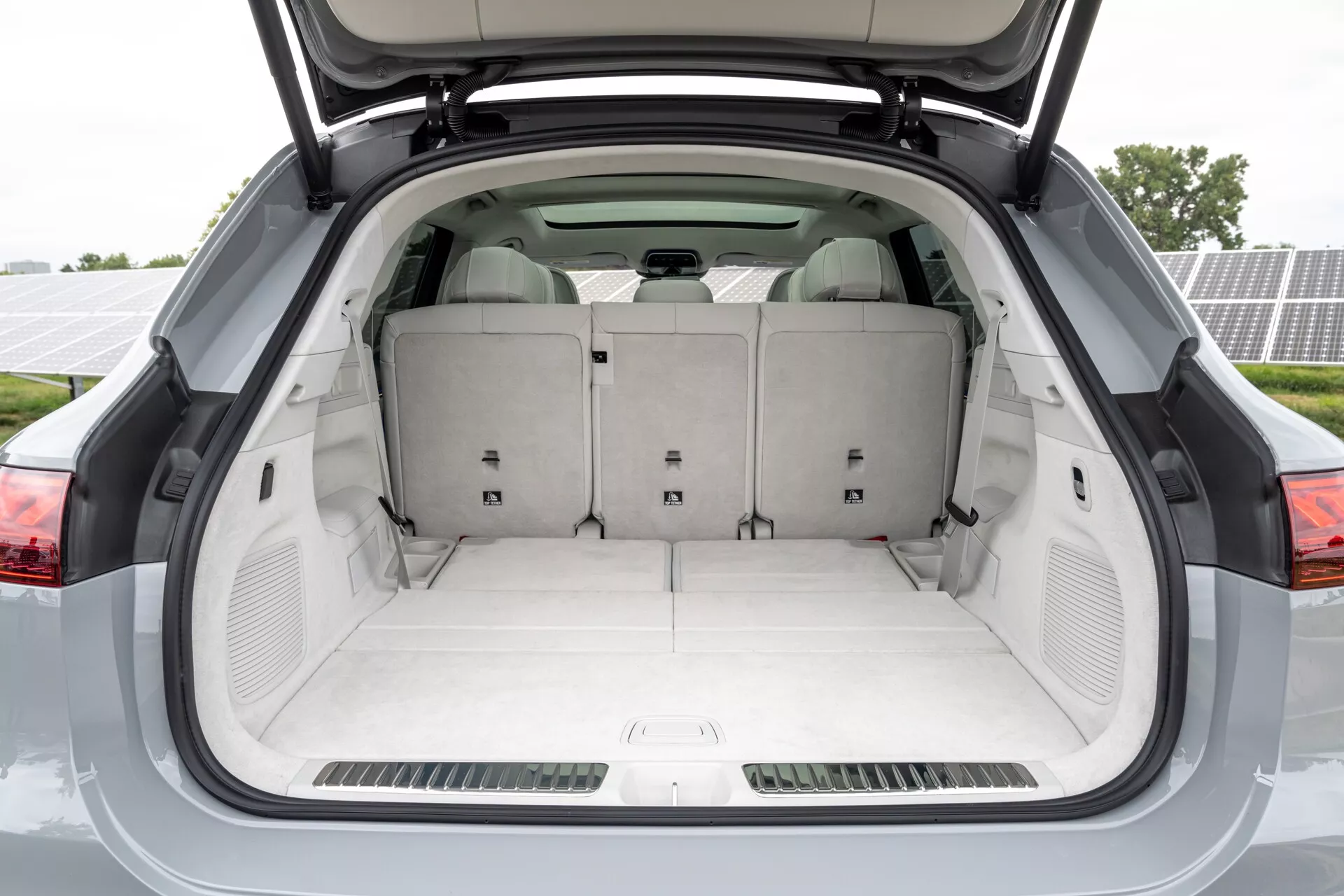 L'image met en évidence l'espace de chargement arrière spacieux du Mercedes EQS SUV 2022, avec une vue directement derrière le hayon ouvert. Le coffre vide montre un large espace de rangement, un sol moquetté et des poches de rangement latérales moulées. Les sièges arrière avec une configuration 60/40 indiquent une polyvalence pour les configurations de chargement et de passagers. La qualité de fabrication premium est évidente, avec des panneaux alignés de manière ordonnée et une cohérence des couleurs. On peut voir des haut-parleurs ou des grilles de ventilation sur les côtés, ainsi que des sangles de traction au plafond et une plaque de protection inférieure pour faciliter le chargement et le déchargement. Cette image reflète des solutions de rangement avancées et des éléments de design pratiques dans un SUV moderne.