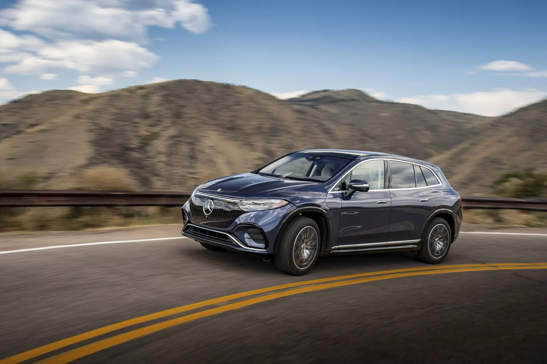 Photo en mouvement d'un SUV Mercedes EQS 2022 moderne roulant sur une route pavée lisse avec une ligne de séparation jaune sous un ciel dégagé. La voiture est vue sous un léger angle de côté avant, mettant en valeur le côté conducteur, y compris la calandre avant, le design des phares, la porte du côté conducteur, le rétroviseur latéral et une partie du pare-brise. Le véhicule est net avec un arrière-plan flou de collines, suggérant le mouvement. Les caractéristiques de design telles que les roues en alliage, les panneaux latéraux sculptés, les lignes distinctives de style et la potentielle technologie ADAS sont visibles.
