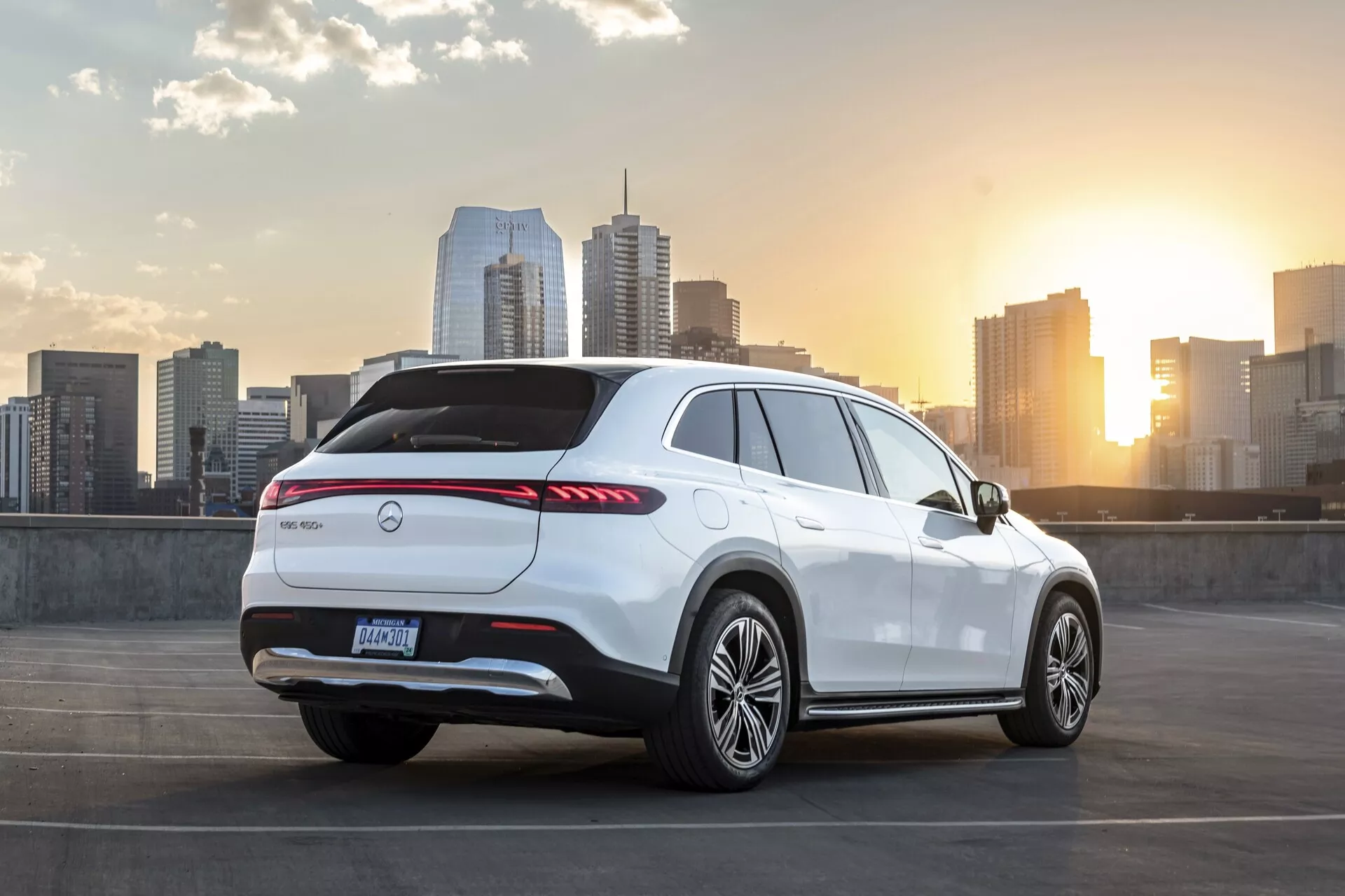 Image du SUV Mercedes EQS 2022 blanc stationné au niveau supérieur d'une structure de parking. Le véhicule est vu sous un angle arrière trois quarts, montrant l'arrière, le côté passager et une partie de la ligne du toit. Il présente des feux arrière LED allongés, l'emblème du fabricant au centre du hayon arrière, un diffuseur arrière, un spoiler arrière, des roues en alliage et une ligne de ceinture haute avec un toit incliné sportif. La scène comprend une skyline de la ville et un coucher de soleil en arrière-plan, soulignant des thèmes de luxe urbain et de mode de vie moderne.