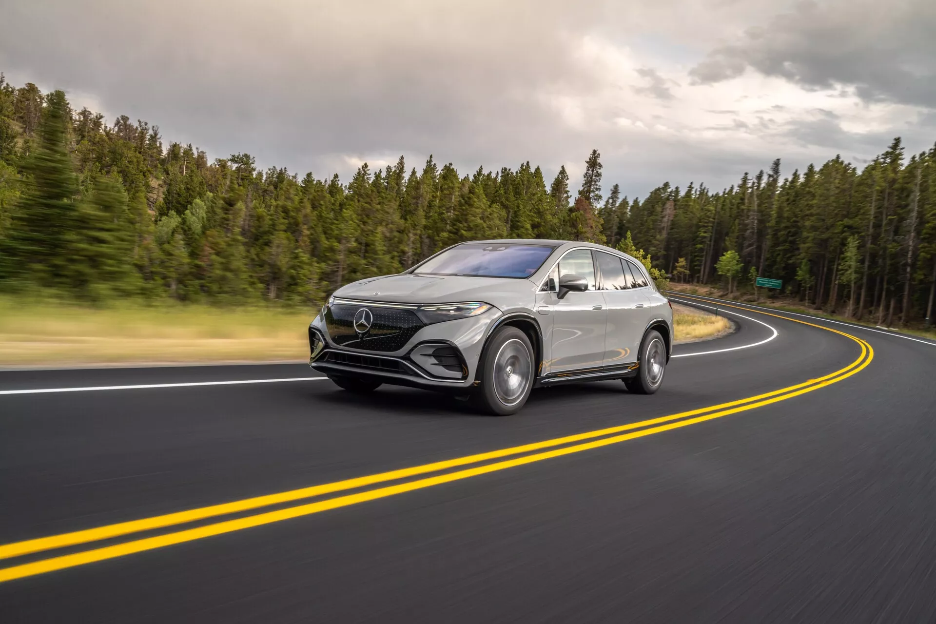 Cette image capture un Mercedes-Benz EQS SUV 2022 en mouvement sous un angle trois-quarts avant avec le design frontal et le profil latéral visibles. Positionné à gauche, la voiture circule sur une route asphaltée bien entretenue avec des doubles lignes jaunes et une courbe vers la droite. Le flou léger sur les roues et un léger flou dû à une vitesse d'obturation plus lente mettent en avant la vitesse et le mouvement dynamique. Les conditions de lumière nuageuses produisent des ombres douces et un éclairage uniforme sur le véhicule. Des caractéristiques technologiques telles que les phares à LED, les feux diurnes, une grande calandre frontale avec un emblème et des rétroviseurs aérodynamiques avec des capteurs potentiels sont visibles. La peinture argentée ou gris clair contraste avec les détails noirs brillants comme la calandre inférieure, les garnitures des passages de roues et les garnitures des fenêtres, mettant en évidence les lignes élégantes, les garde-boue incurvés et la ligne de toit ressemblant à un coupé. Adapté pour les catégories automobile, technologie, photographie de mouvement et véhicules de luxe.