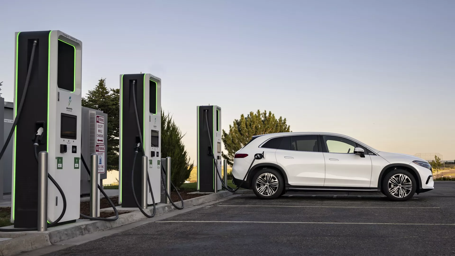 Un véhicule électrique (EV) moderne et élégant, le Mercedes EQS-SUV 2022, montré à une station de recharge dans des conditions d'éclairage doux, soit en fin d'après-midi, soit tôt le matin. Le profil latéral complet révèle des lignes aérodynamiques, des roues distinctives, des vitres teintées et un habitacle bien défini. Le véhicule est connecté à une station de recharge moderne, soulignant l'accent mis sur les transports durables et l'infrastructure des véhicules électriques.