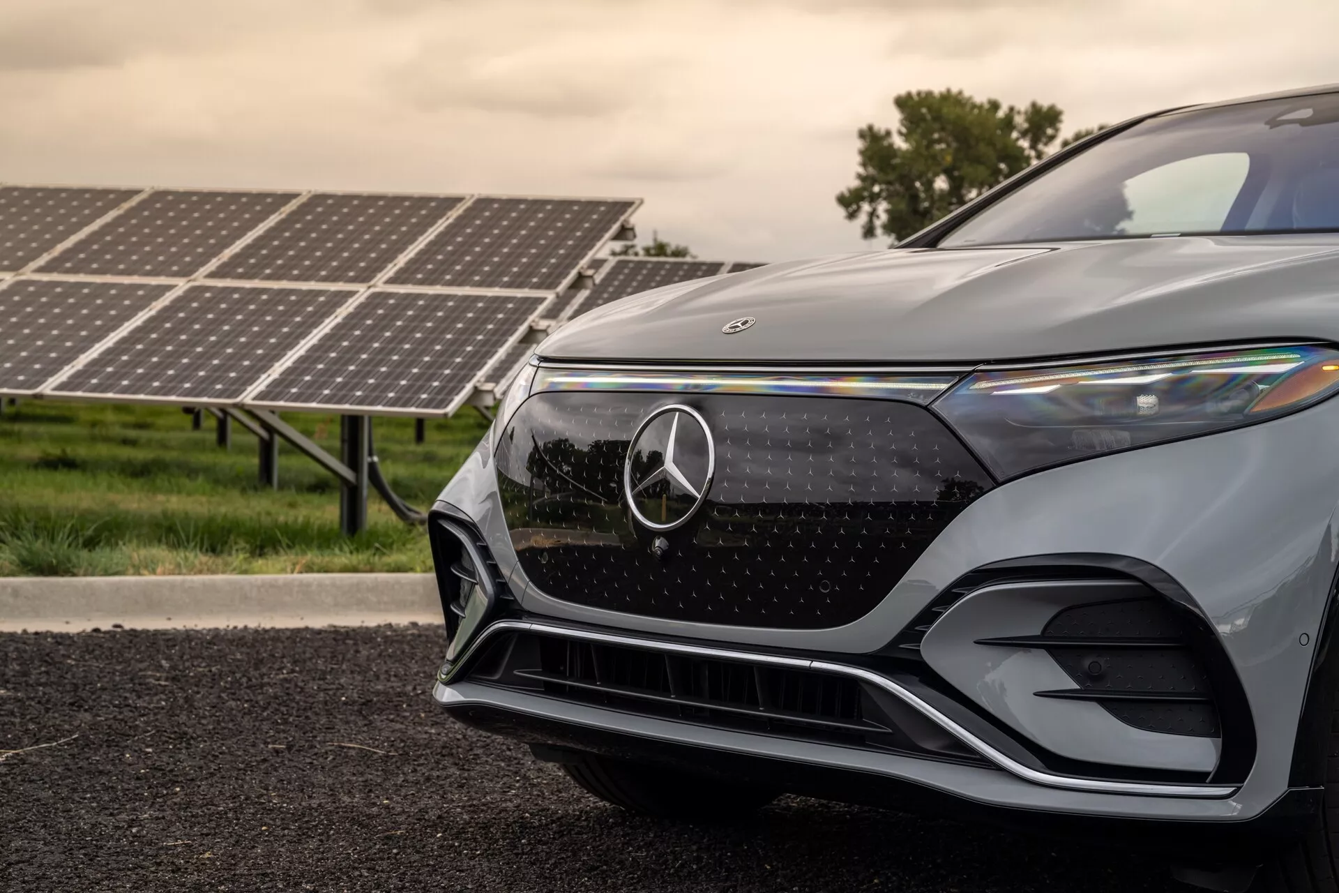 Gros plan du quart avant gauche d'un SUV Mercedes-Benz EQS 2022 moderne, pris d'un angle bas. L'image met en valeur la grille avec l'emblème Mercedes-Benz, le phare LED angulaire et élégant, le pare-chocs avant aérodynamique et la finition de peinture métallique. Les panneaux solaires en arrière-plan suggèrent la nature électrique ou hybride de la voiture, soulignant ses caractéristiques technologiques avancées et respectueuses de l'environnement.
