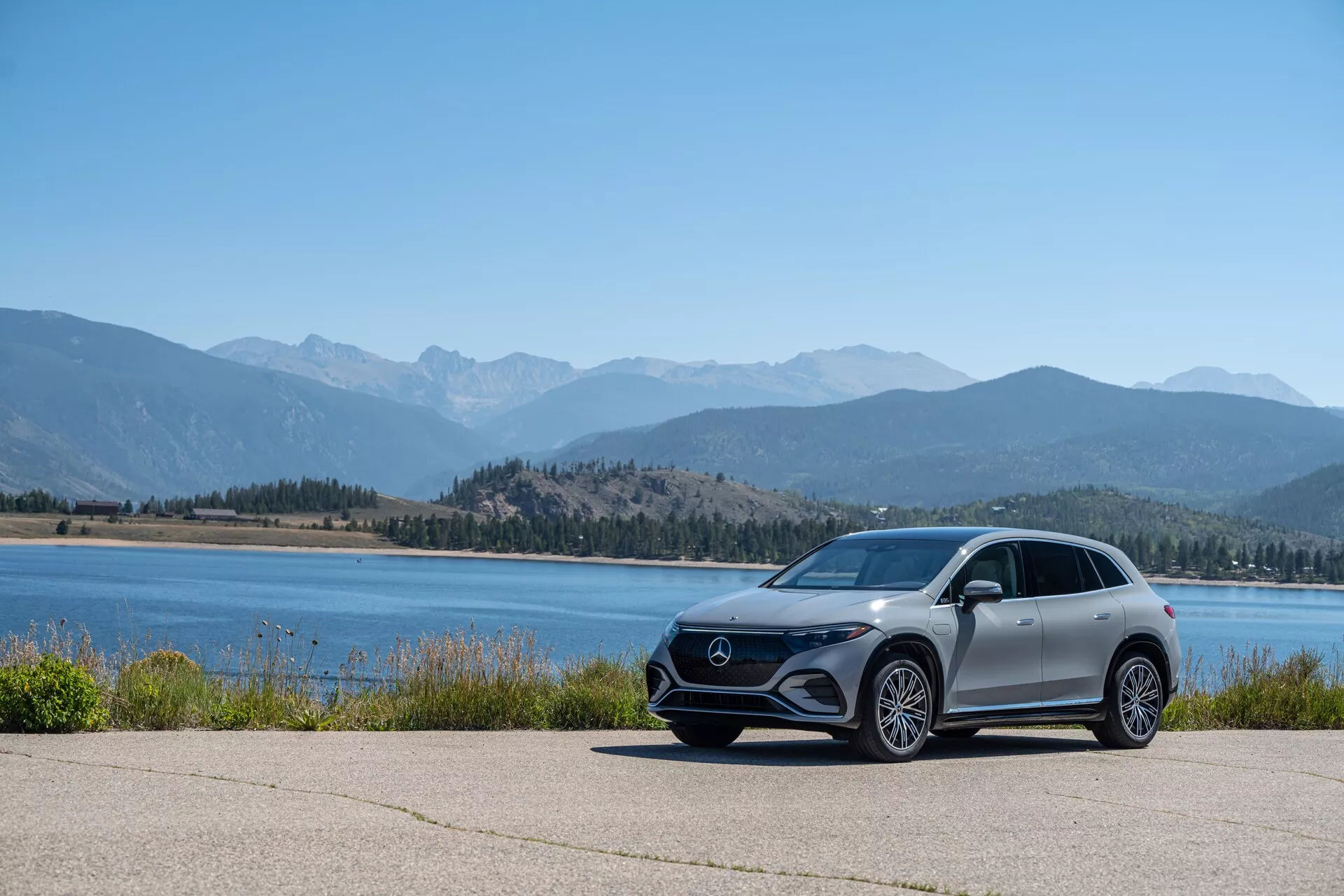 L'image montre un Mercedes EQS SUV 2022 moderne, garé sur une zone pavée avec un panorama pittoresque de plans d'eau et de chaînes de montagnes, prise d'un angle bas pour mettre en valeur sa posture et ses lignes de design. La voiture est vue de profil latéral avec une visibilité de la grille avant, des phares LED angulaires, de grandes roues en alliage, des rétroviseurs latéraux aérodynamiques et une finition de peinture métallique.