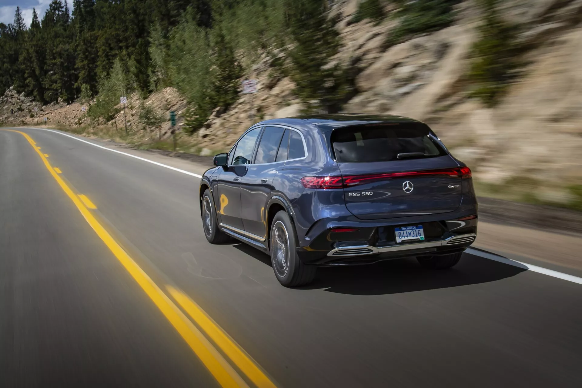 Mercedes-Benz EQS SUV 2022 moderne et luxueux capturé en mouvement sur une route pavée, montrant un design élégant avec peinture sombre, feux arrière à LED horizontaux, spoiler monté sur le toit, double échappement et jantes en alliage multi-branches. Une technique de filé à grande vitesse souligne le mouvement du véhicule à travers un paysage naturel et montagneux.