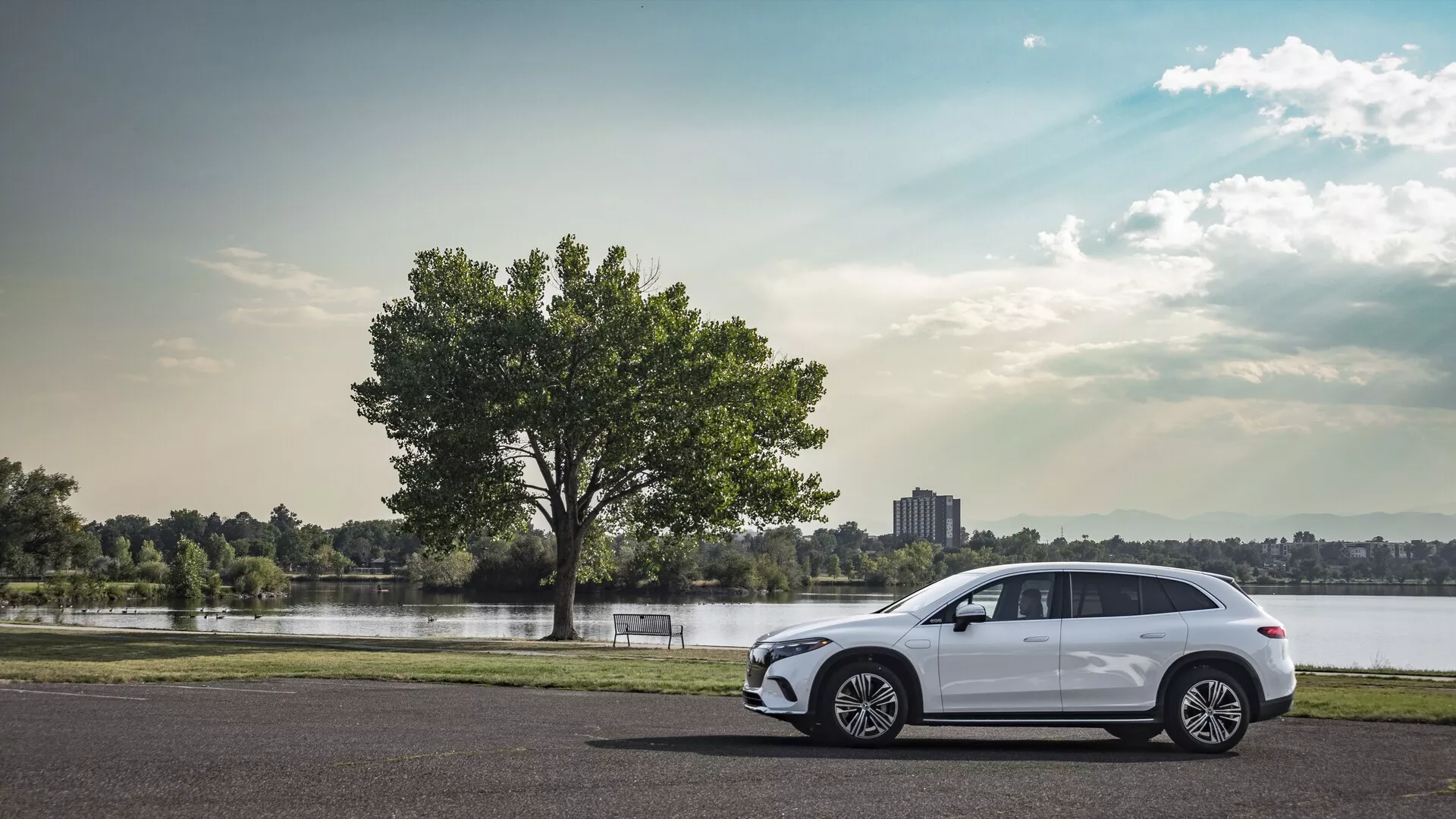 Vue de profil latéral du moderne Mercedes EQS SUV 2022 stationné sur de l'asphalte, avec des roues en alliage multi-branches, des lignes aérodynamiques élégantes, quatre portes, fenêtres teintées avec rétroviseur latéral à clignotant intégré, ligne de toit sportive avec barres de toit et feux arrière à LED partiellement visibles, le tout sur un arrière-plan serein comprenant un lac ou une rivière, un arbre, de l'herbe et un ciel partiellement nuageux.