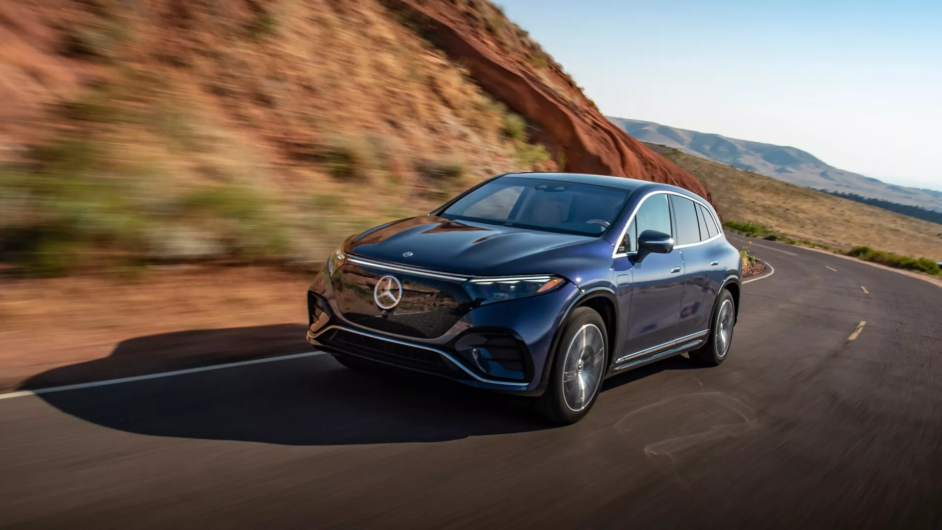 Prise de vue dynamique du Mercedes EQS-SUV 2022 capturée sous un angle de trois quarts avant gauche montrant le véhicule en mouvement. Les caractéristiques incluent le côté gauche élégant de la voiture, le capot avant, les phares avec technologie LED avancée, la grille, le pare-chocs avant et les roues en alliage. Le design aérodynamique de la carrosserie indique luxe et ingénierie avancée, mettant en valeur les performances et l'expérience de conduite. L'image émet un sentiment de liberté et de performance sur des routes dégagées entourées de nature.