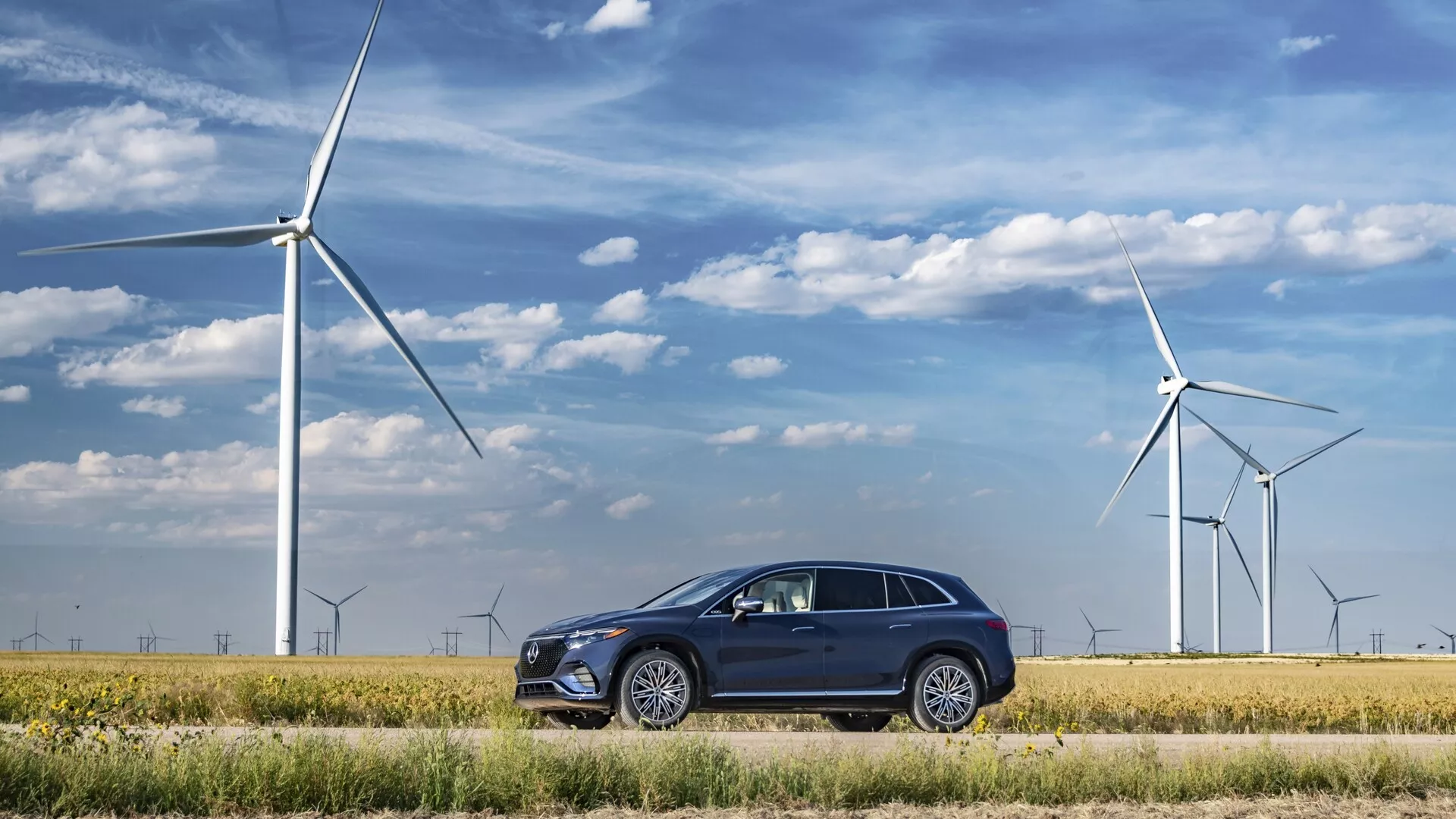 Mercedes EQS-SUV 2022 stationné dans un paysage écologique avec des éoliennes en arrière-plan, montrant un design moderne et élégant avec une carrosserie sombre, des jantes en alliage multibrins, des poignées de porte affleurantes, des garnitures chromées autour des fenêtres, des phares LED angulaires et des rétroviseurs avec clignotants intégrés.