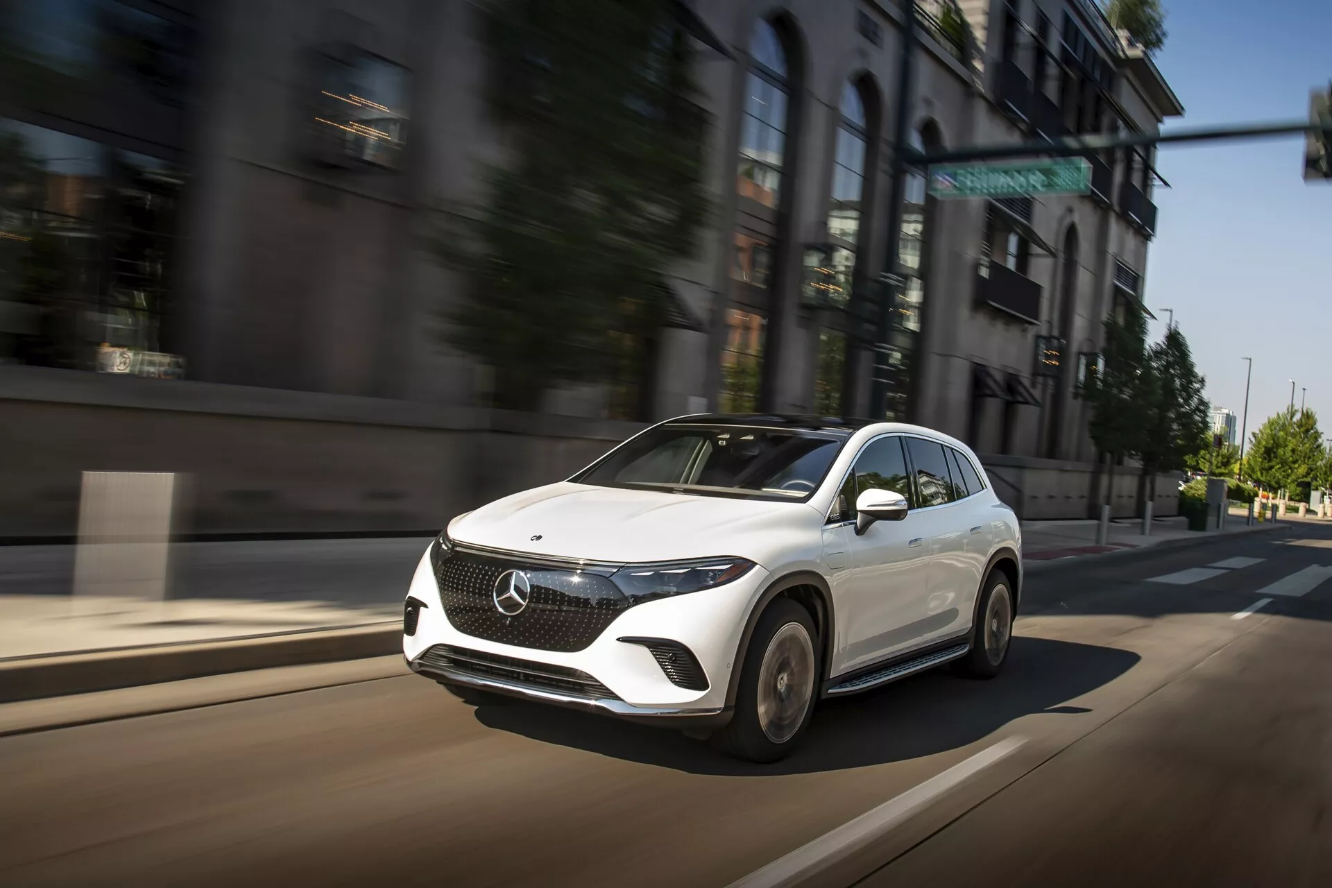 L'image capture une Mercedes EQS-SUV 2022 en mouvement sur une rue urbaine, prise sous un angle de trois quarts de face, mettant en valeur sa face avant, son capot sculpté, ses élégants phares LED angulaires et une partie du design des roues. La couleur blanche du véhicule contraste avec l'arrière-plan urbain flou, accentuant sa vitesse et sa présence dynamique. Le grand emblème sur la grille, le rétroviseur latéral aérodynamique et la configuration avancée des phares suggèrent ses caractéristiques modernes, luxueuses et technologiquement avancées.