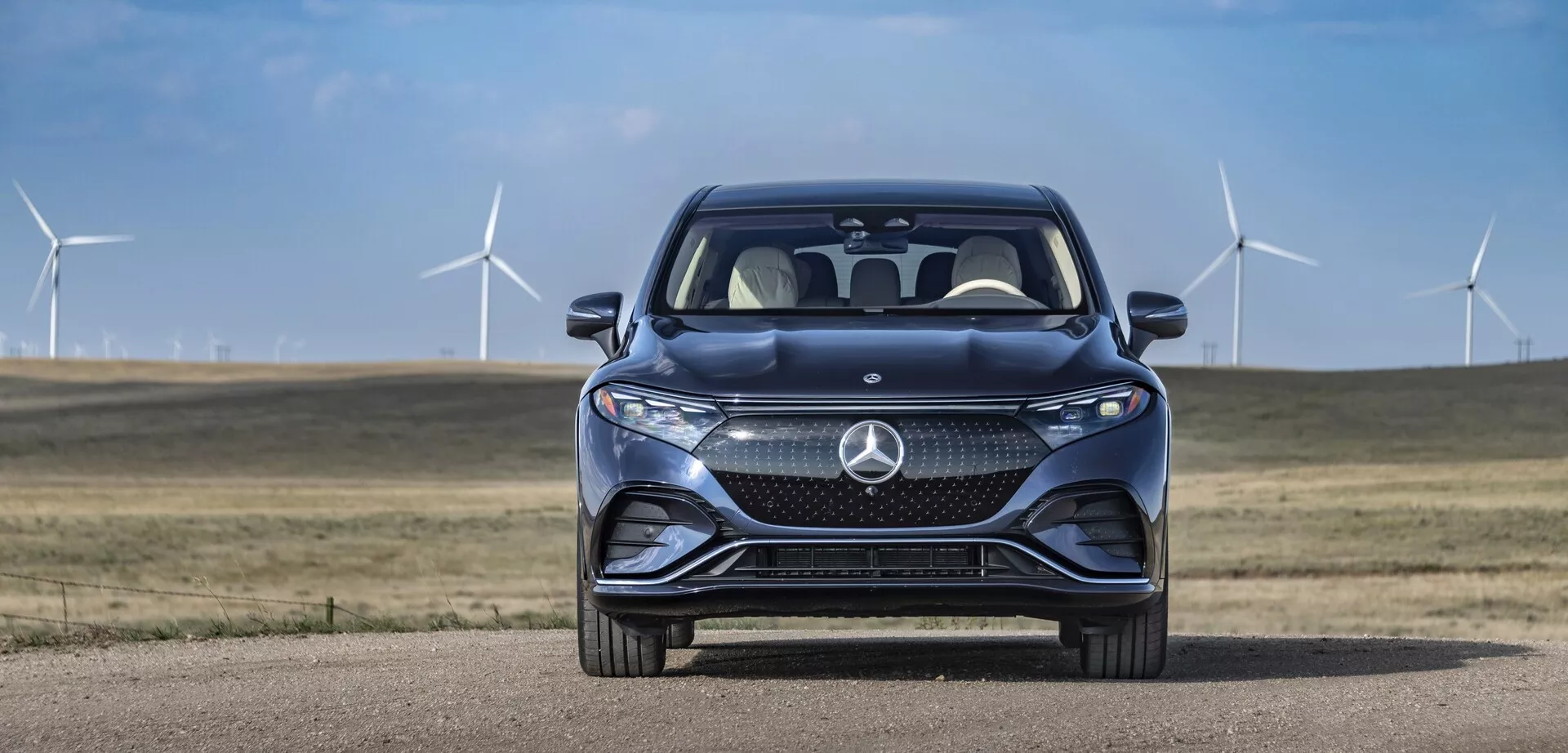 Vue de face du Mercedes EQS-SUV 2022 contre un ciel dégagé et des éoliennes en arrière-plan. La grille proéminente du véhicule présente un grand emblème stylisé, avec des motifs en maille complexes probablement abritant des capteurs pour les systèmes d'assistance à la conduite. Des phares LED horizontaux et élégants avec des feux de jour intégrés sont visibles. Le pare-chocs avant sport comprend de grandes prises d'air, et les lignes de contour du capot s'inclinent vers l'avant pour améliorer l'aérodynamisme. La vue partielle du quart avant révèle une partie des arches de roue, indicatif d'un design premium. Le pare-brise réfléchissant et le rétroviseur latéral côté passager partiellement visible suggèrent des fonctionnalités avancées, comme les clignotants et la surveillance des angles morts. Les systèmes avancés d'assistance à la conduite et les indices de durabilité sont suggérés par l'environnement.