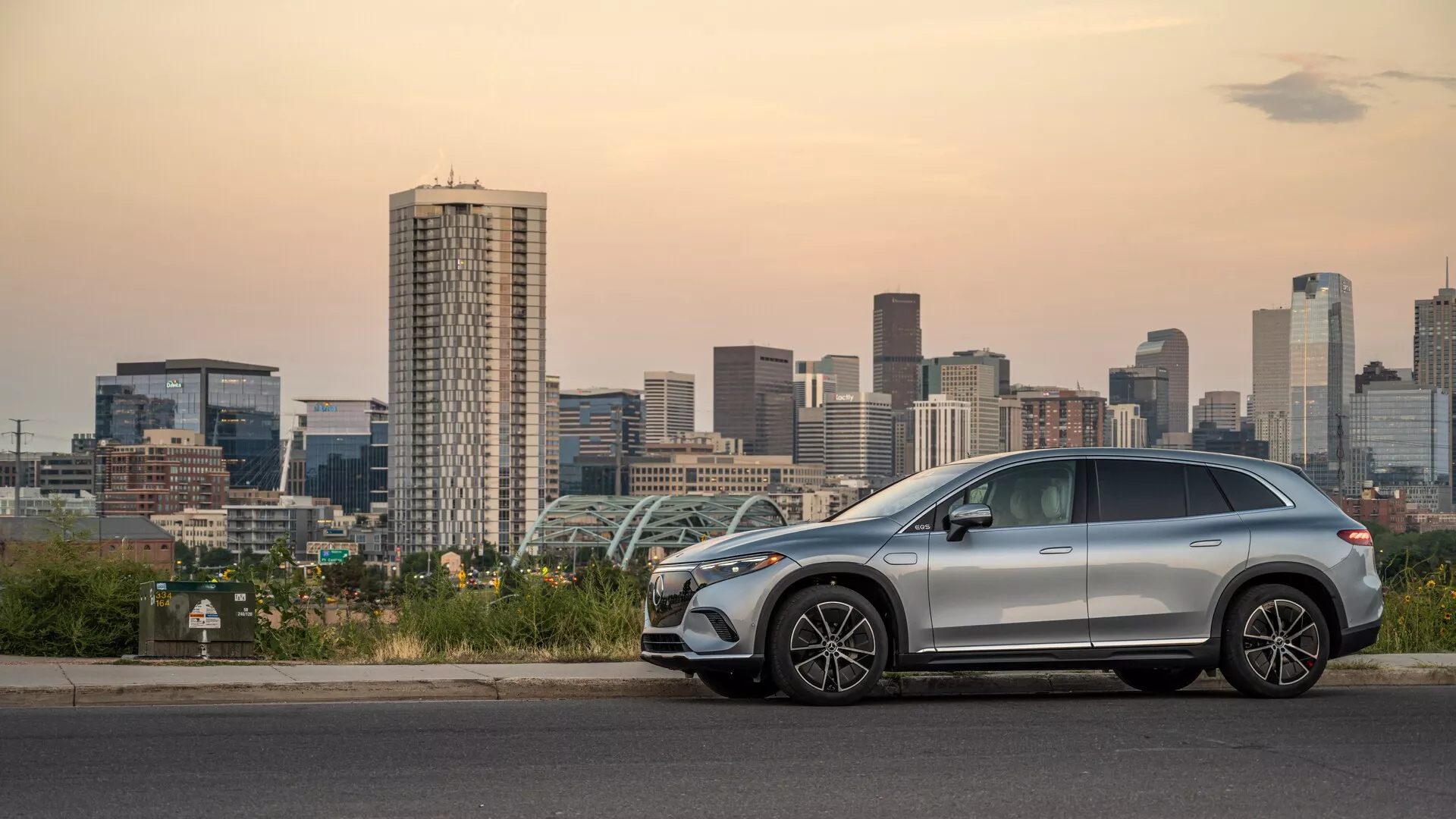 Vue latérale d'un SUV Mercedes EQS moderne et de taille moyenne stationné près d'une ligne d'horizon urbaine au crépuscule, montrant un design aérodynamique élégant, des roues en alliage, des barres de toit, un possible toit ouvrant et des caractéristiques avancées telles que des rétroviseurs latéraux avec clignotants intégrés et une entrée sans clé.