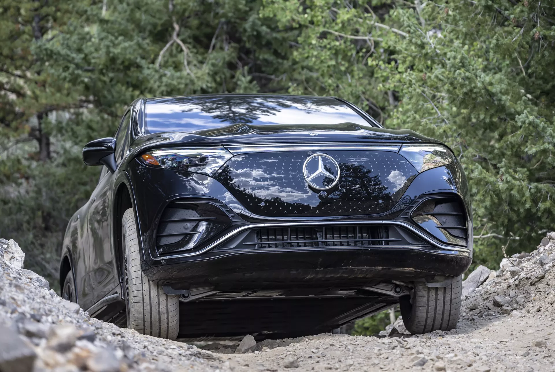 Prise de vue en contre-plongée d'un Mercedes-Benz EQS SUV 2022 noir, mettant en valeur son capot sculpté, son grand emblème étoilé central, sa calandre complexe, ses élégants phares à LED et ses prises d'air inférieures. Capturé dans un environnement naturel en plein air, le véhicule, situé sur une pente, montre sa garde au sol élevée et son potentiel tous terrains. Le véhicule est propre avec quelques taches de saleté, soulignant son utilisation légère hors route et son design automobile avancé.