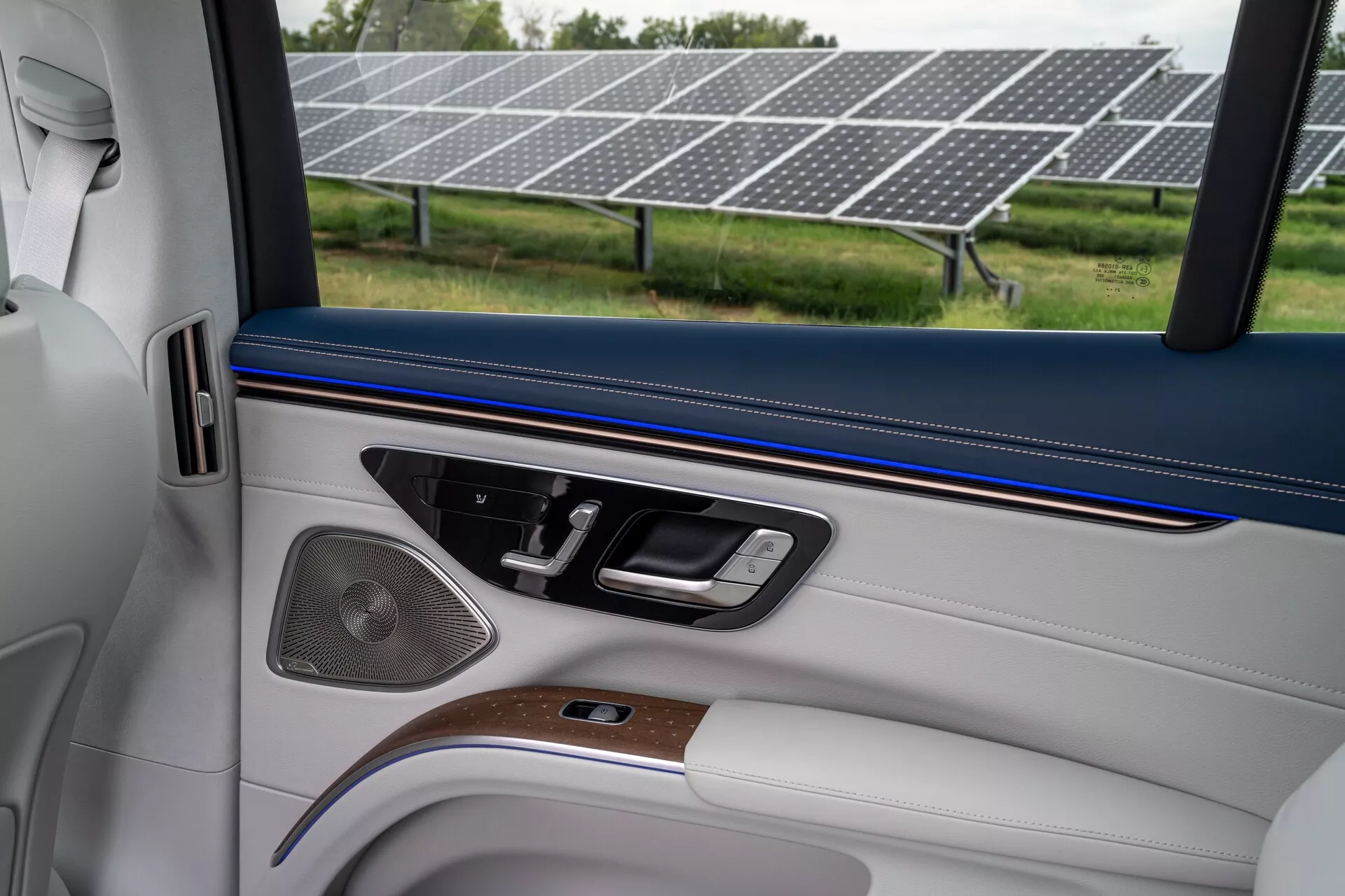 Intérieur d'un Mercedes EQS SUV 2022, axé sur la porte côté passager. La porte présente un décor bicolore avec une garniture supérieure bleu foncé et une section inférieure blanche ou gris clair. Elle comporte une pièce en bois et des accents métalliques autour de la poignée. Les commandes des fenêtres, des miroirs et des serrures sont visibles, ainsi qu'une grille d'enceinte et une poche de porte.