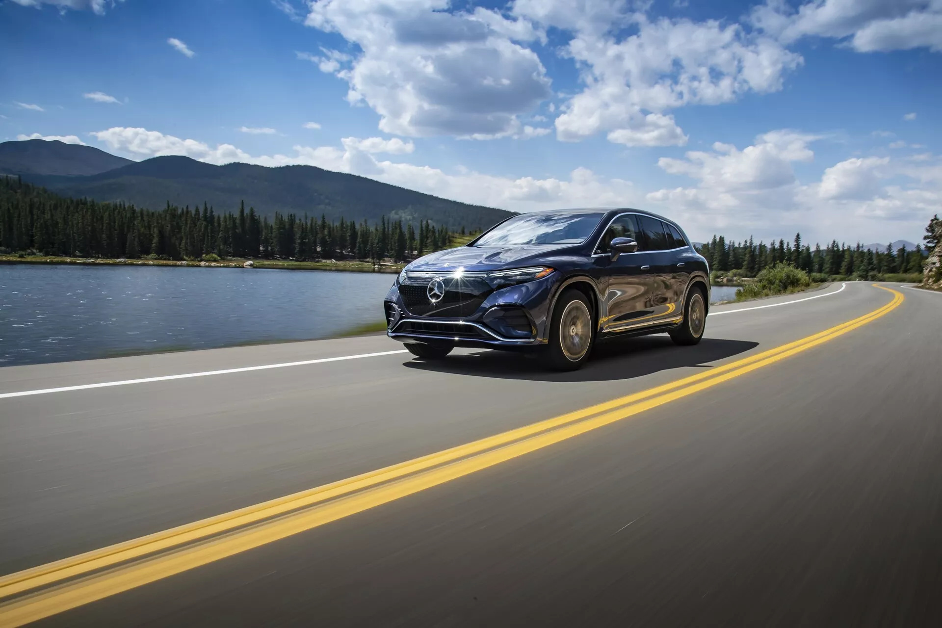 SUV Mercedes EQS 2022 roulant sur une route asphaltée avec des lignes jaunes doubles sous un ciel partiellement nuageux en journée. Le véhicule est capturé sous un angle de trois quarts avant bas, mettant en évidence la calandre avant proéminente avec un grand emblème, les phares à LED, les lignes élégantes du capot, les roues en alliage et un rétroviseur latéral avec des clignotants intégrés possibles. L'arrière-plan présente un paysage serein avec un plan d'eau, des montagnes et un horizon dégagé, soulignant le sentiment de voyage et d'aventure. Le mouvement de la voiture est mis en valeur par un léger flou de mouvement.