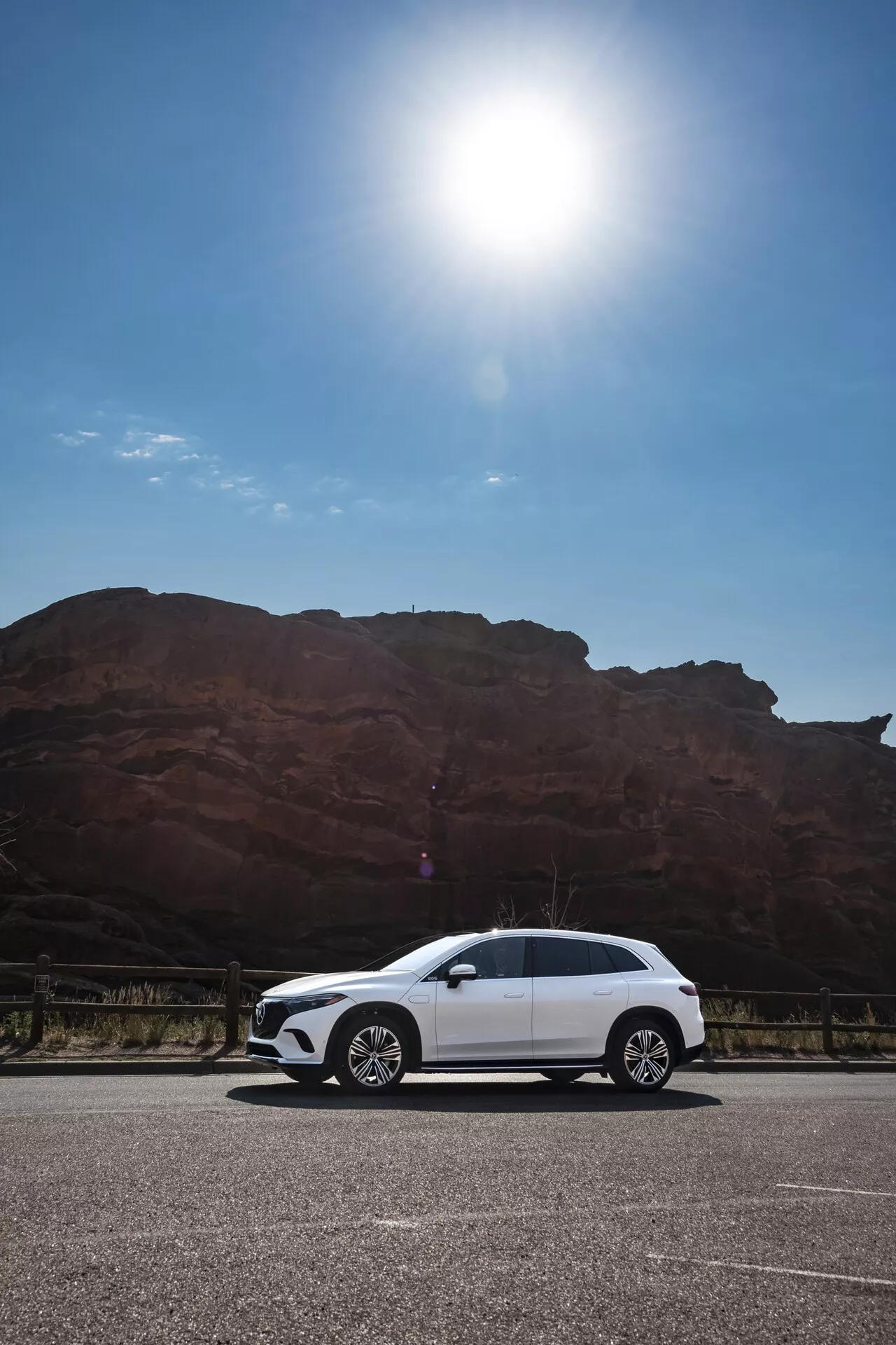 Image diurne d'un Mercedes EQS SUV 2022 blanc stationné sur une zone pavée devant une grande formation rocheuse rouge-brun, prise d'un angle bas montrant le profil latéral de la voiture y compris les poignées de porte, rétroviseurs latéraux, jantes et contours de la carrosserie sous un ciel dégagé avec une éblouissante lumière du soleil dans le coin supérieur gauche.