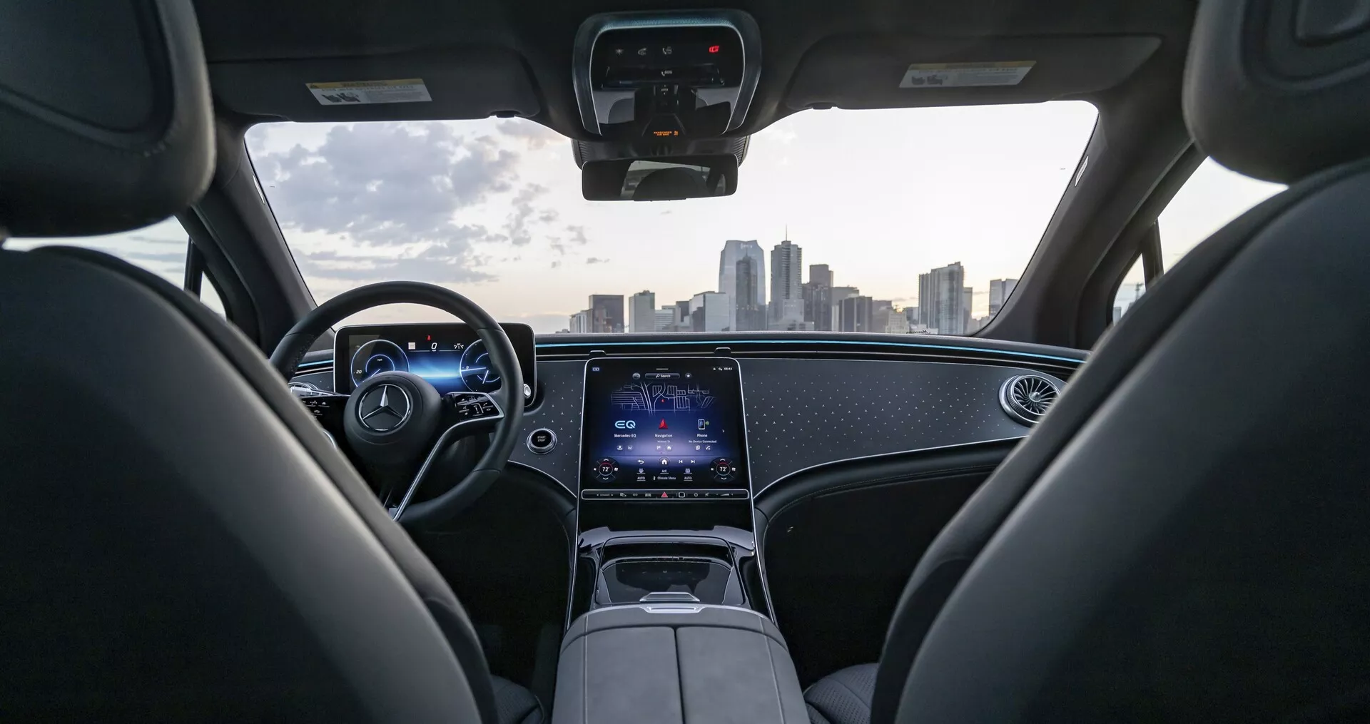 Vue intérieure d'une Mercedes EQS-SUV 2022 depuis la perspective du siège arrière, montrant un tableau de bord moderne avec éclairage d'ambiance, un tableau de bord numérique et un volant en cuir avec commandes intégrées. Un grand écran tactile domine la console centrale pour les fonctions d'infodivertissement et de connectivité. Le pare-brise offre une vue claire et dégagée de la ligne d'horizon de la ville, mettant en valeur la technologie avancée et le design luxueux de la voiture.