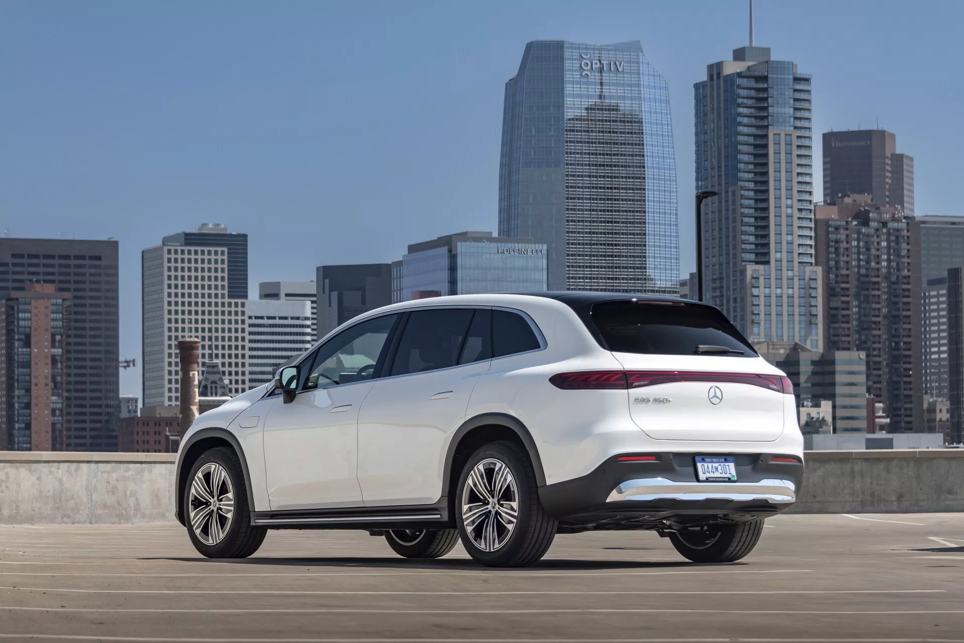Vue arrière trois quarts d'un Mercedes EQS-SUV 2022 blanc depuis un angle légèrement élevé, montrant la partie supérieure de la section arrière, le profil latéral et des caractéristiques de design distinctives telles que les feux arrière LED élégants, le hayon, le pare-chocs arrière et les grandes roues en alliage multi-branches. Le SUV est garé dans un parking en plein air avec une ligne d'horizon urbaine en arrière-plan, mettant en valeur son design moderne et haut de gamme avec d'éventuelles fonctionnalités avancées typiques des modèles de luxe.