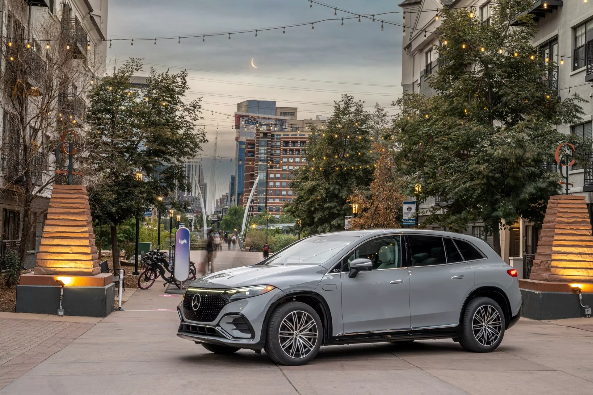 SUV moderne en milieu urbain lors de l'aube ou du crépuscule, prise en contre-plongée du quart avant mettant en valeur la calandre avant, les phares, le capot et une partie du côté conducteur. Composition centrée avec des pots de fleurs et des réverbères chauds ajoutant de la profondeur. Caractéristiques de luxe distinctives incluent des roues en alliage à plusieurs rayons, des phares à LED, une calandre avec un grand emblème, des clignotants intégrés aux rétroviseurs, des lignes de design élégantes et une peinture métallique.