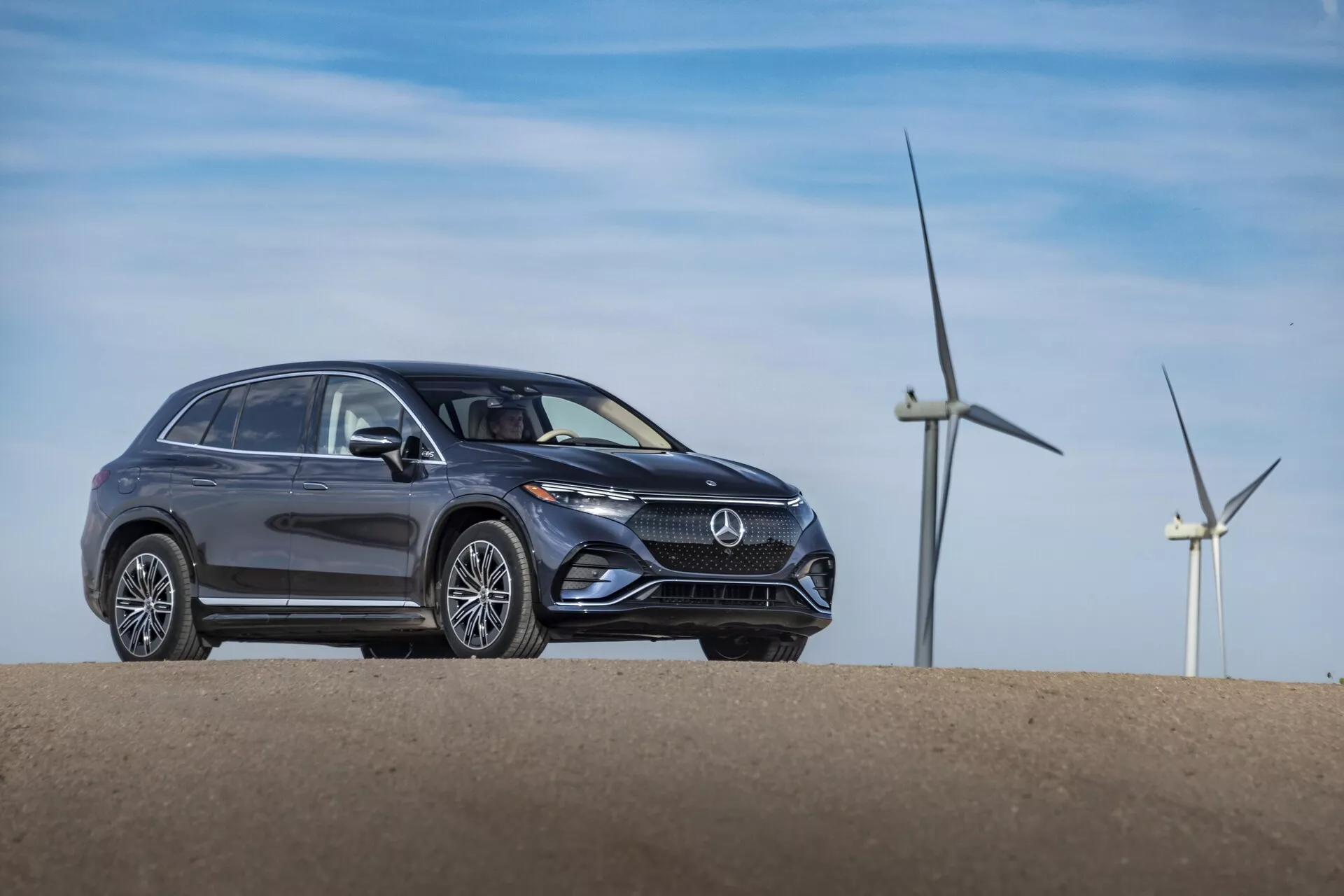 Vue de trois quarts avant d'un Mercedes EQS-SUV 2022 gris positionné sur une petite colline, présentant un design moderne avec des lignes de carrosserie élégantes, des formes distinctives des phares, une calandre avant proéminente avec un emblème de logo, des roues stationnaires et des jantes en alliage. Les éoliennes en arrière-plan indiquent peut-être un parc éolien ou une zone rurale, évoquant potentiellement des attributs écologiques du véhicule.