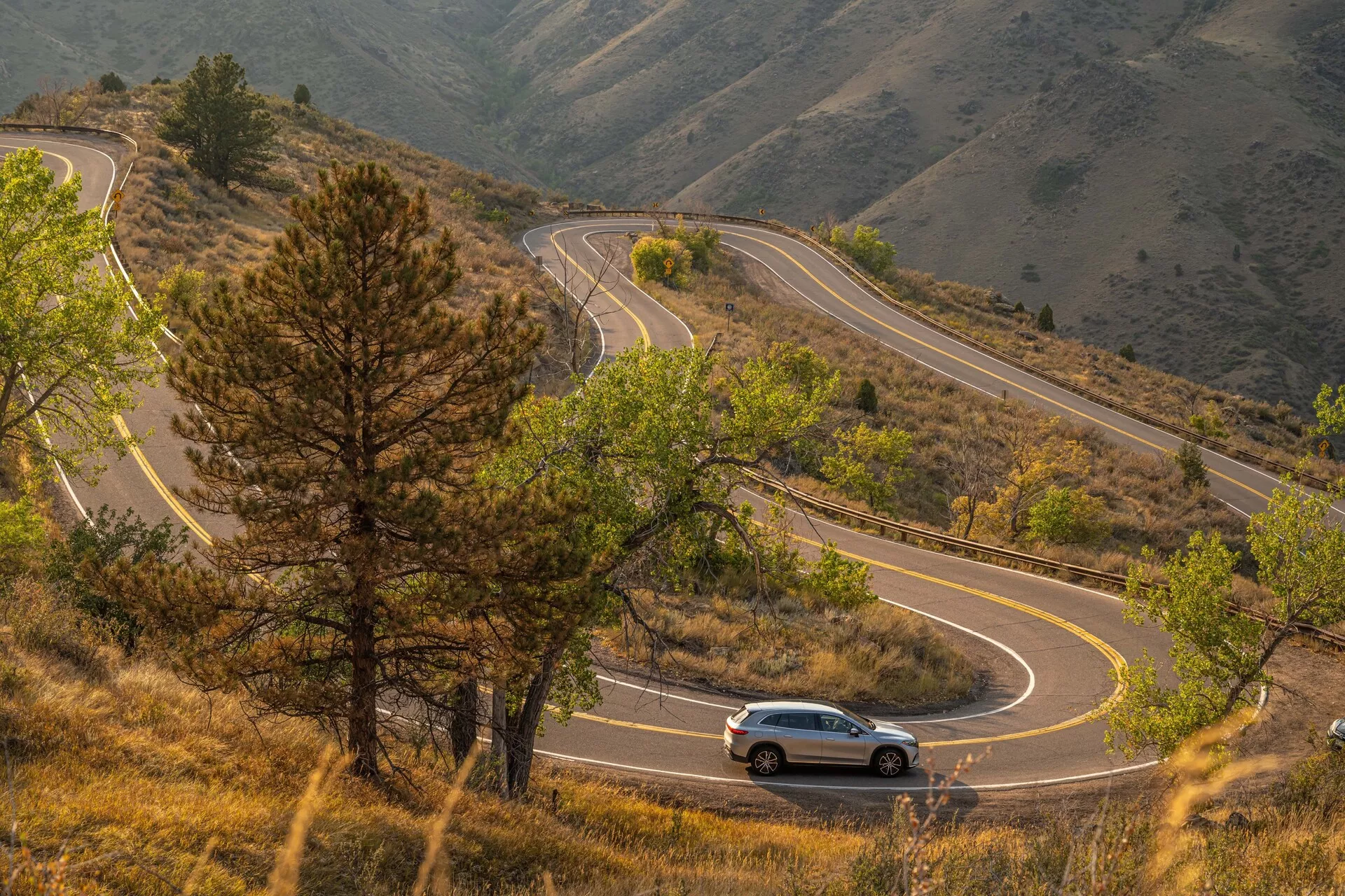 Vue en plongée d'un SUV Mercedes EQS 2022 naviguant sur une route de montagne sinueuse entourée d'un paysage vallonné et boisé. La voiture est centrée dans l'image, offrant une perspective latérale et légèrement arrière, mettant en avant ses lignes de design, sa forme aérodynamique et ses phares et feux arrière visibles.