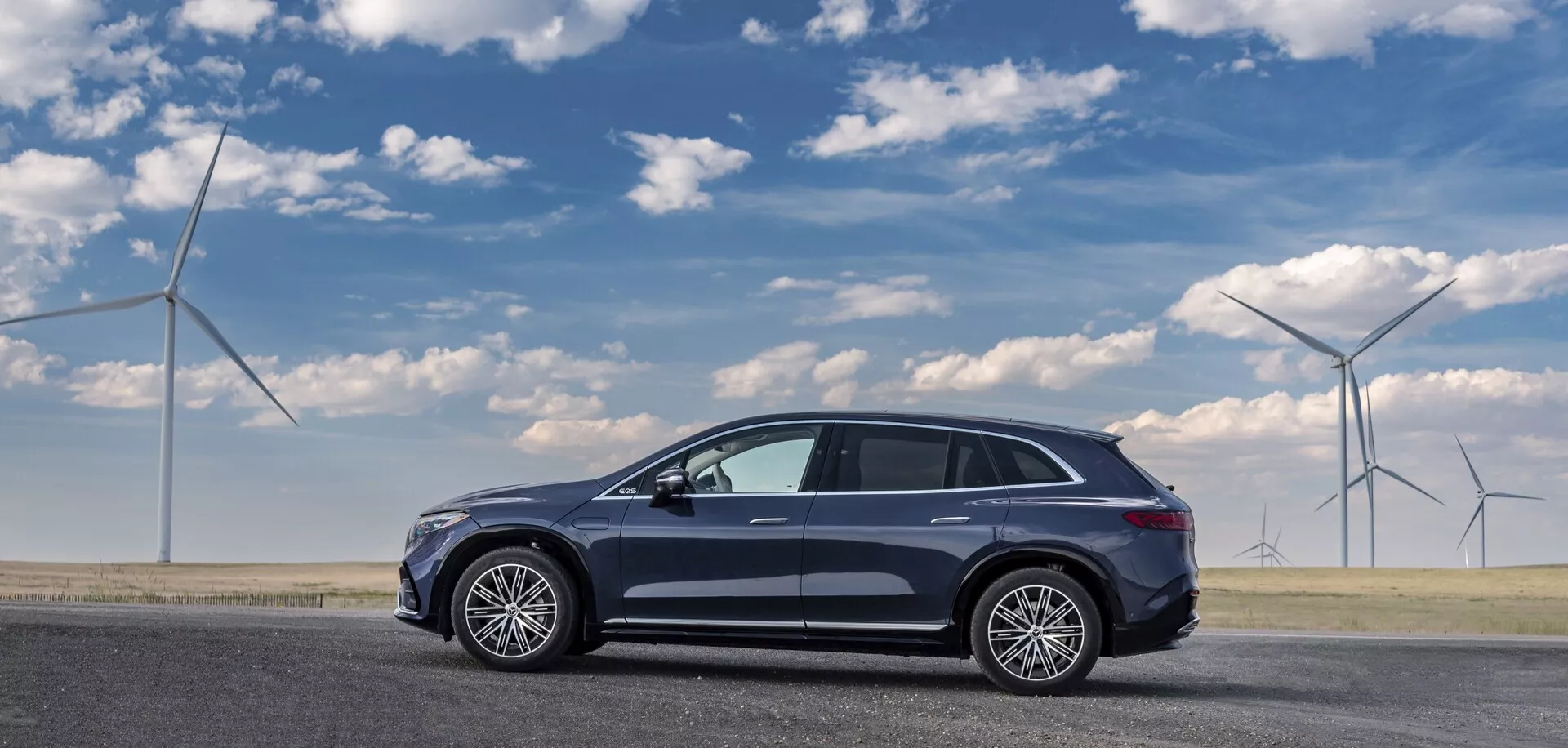 Vue de côté d'une Mercedes EQS-SUV 2022 de couleur sombre garée sur une surface routière plate, capturée du côté passager. Le crossover SUV moderne aérodynamique et élégant présente cinq portes, de grandes roues en alliage à plusieurs branches, un toit ouvrant panoramique, une antenne de toit et des rétroviseurs latéraux avec clignotants intégrés, avec une finition métallique. Le profil de la voiture suggère une position élevée typique des crossovers. Les éoliennes en arrière-plan suggèrent des thèmes écologiques.