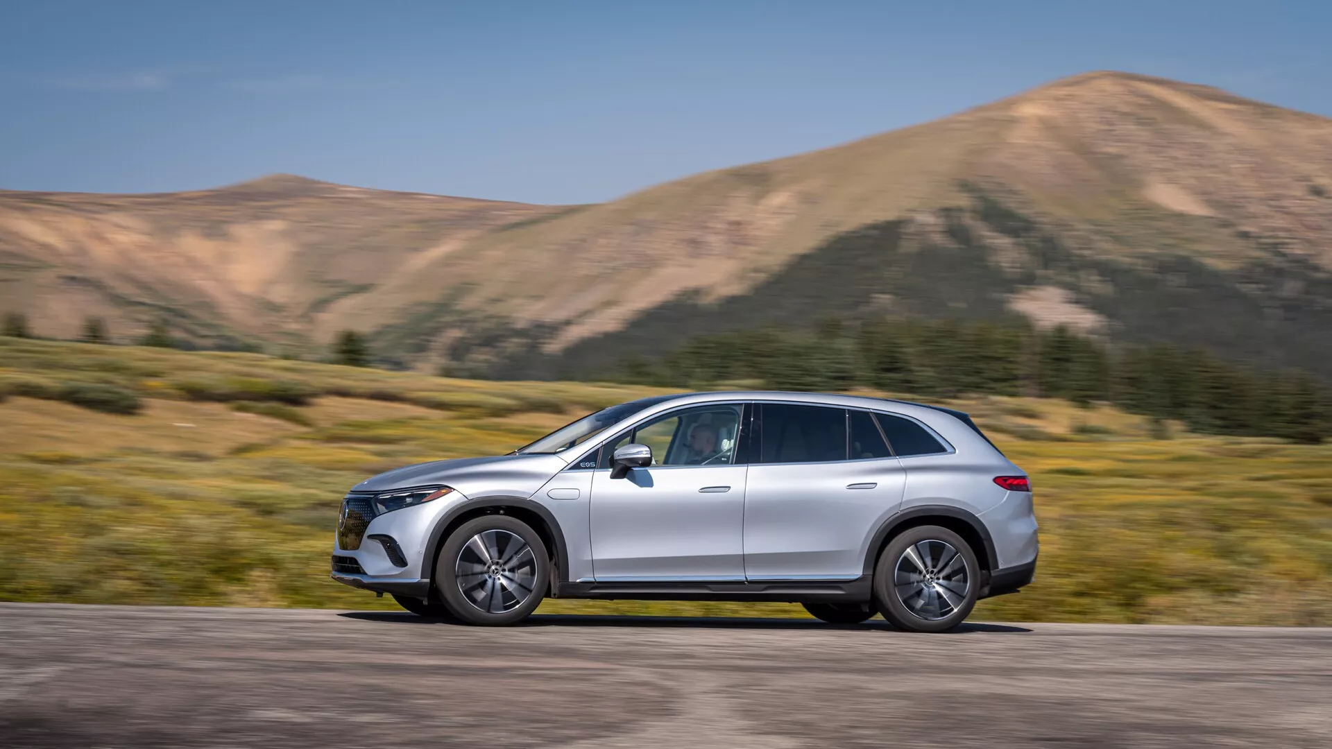 Mercedes EQS-SUV 2022 capturé en vue de profil, occupant la position centrale du cadre avec un mouvement évident par un arrière-plan légèrement flou. Le SUV de grande taille arbore un design élégant et profilé, des rétroviseurs latéraux aérodynamiques, des jantes en alliage, des barres de toit, des garnitures chromées et une peinture métallique. Des éléments technologiques avancés sont visibles, notamment des feux de jour à LED et des poignées de porte affleurantes suggérant l'entrée sans clé. Un décor montagneux renforce son attrait dynamique et aventureux, idéal pour des annonces automobiles et des contextes de style de vie.