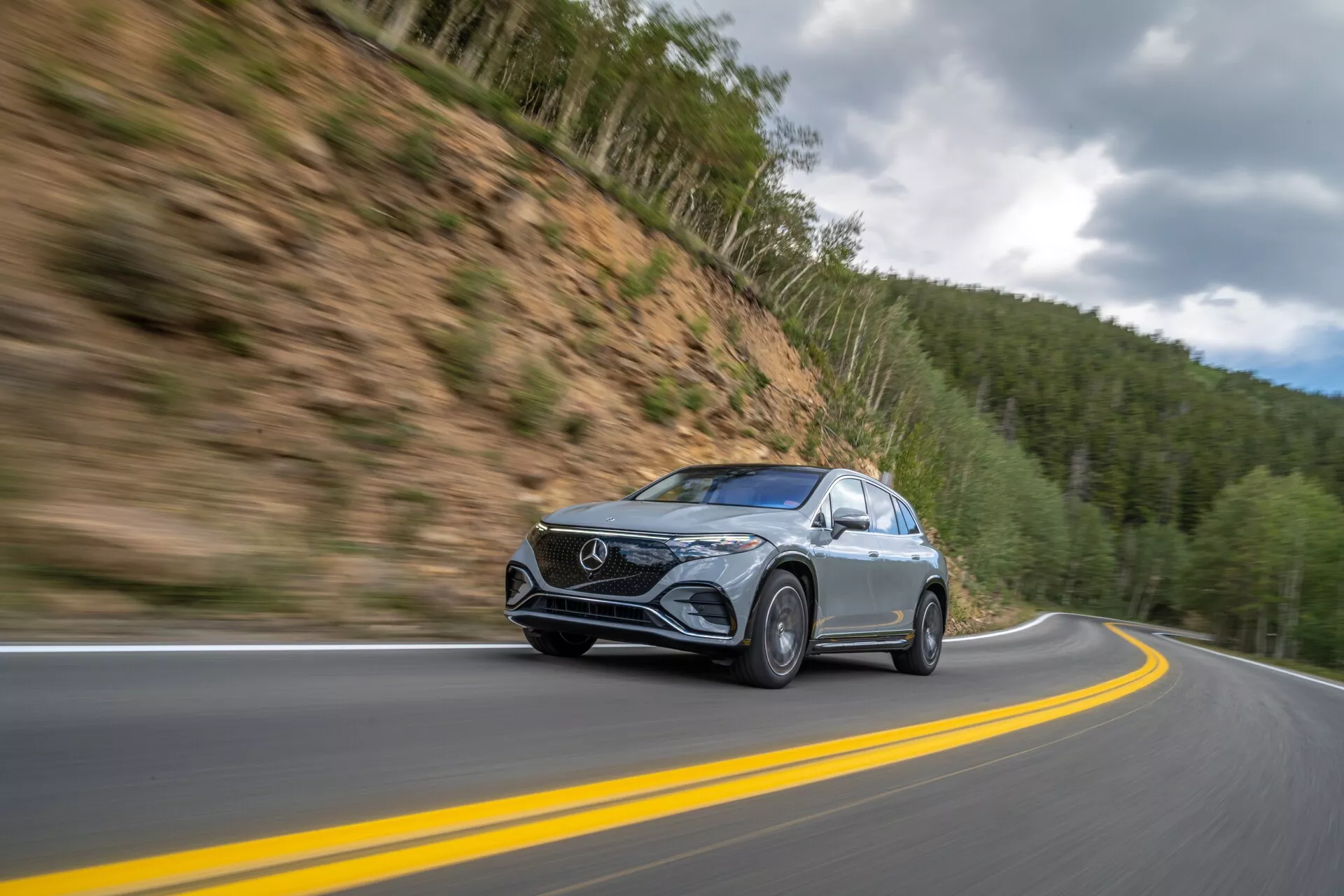 Prise de vue dynamique d'un Mercedes EQS-SUV 2022 en mouvement sur une route de montagne, capturée sous un angle bas et légèrement frontal. La voiture, située à gauche du centre, se déplace le long d'une route à deux voies sinueuses avec un arrière-plan flou indiquant une vitesse élevée. Doté de phares à LED élégants, d'une calandre avant distincte et d'une peinture métallique, le véhicule se détache sur un arrière-plan montagneux boisé sous un ciel nuageux. Des détails comme les roues floues en mouvement et les rétroviseurs latéraux clairs avec des clignotants intégrés éventuels renforcent l'attrait technologique.