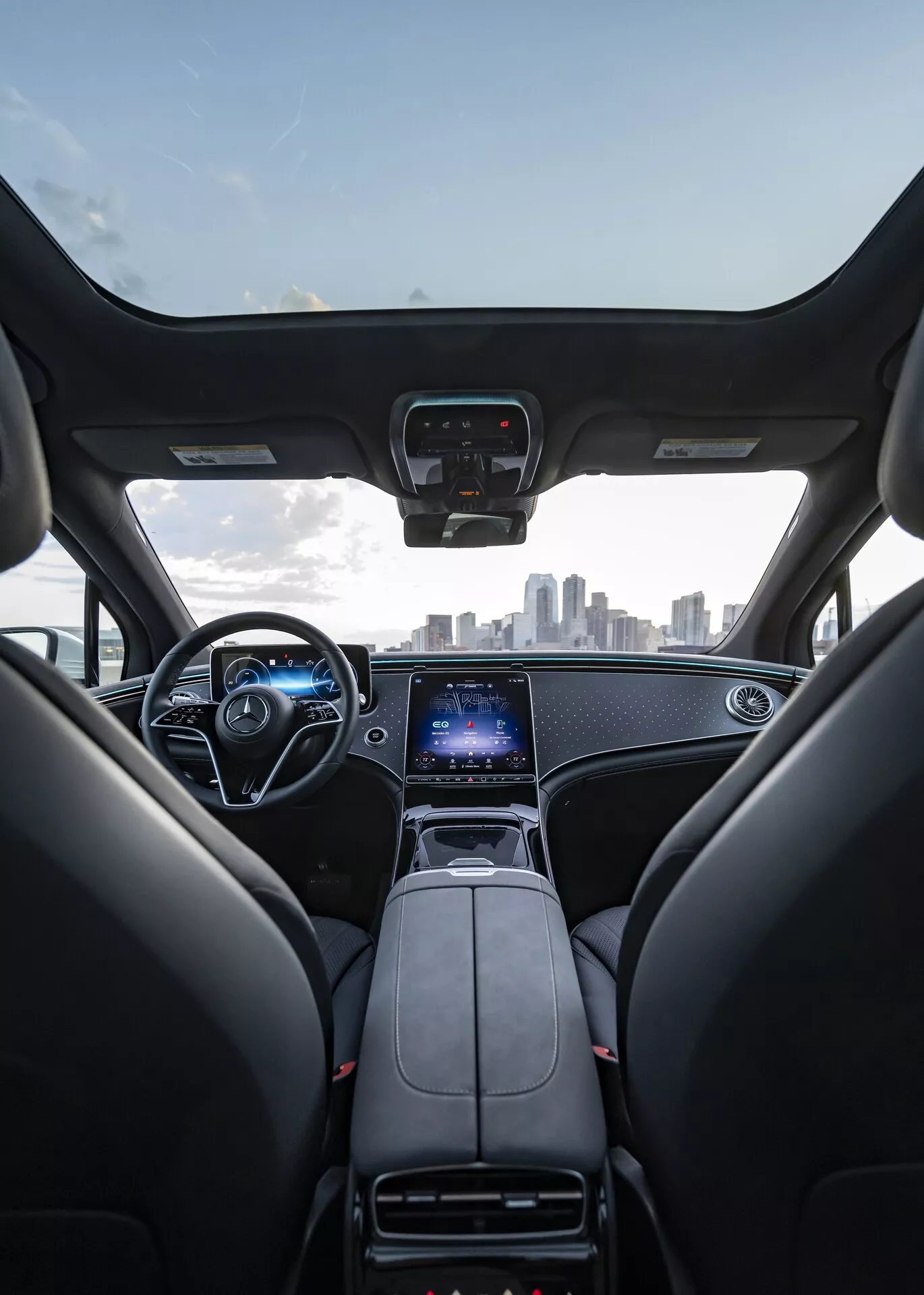 Vue intérieure du Mercedes EQS SUV 2022 depuis la perspective du siège arrière, montrant les sièges avant, le tableau de bord, le volant et le pare-brise. Les sièges avant sont en cuir noir avec appuie-têtes, et le tableau de bord présente un design élégant avec des accents métalliques. La console centrale abrite un levier de vitesse, des porte-gobelets et un grand écran tactile vertical pour le système d'info-divertissement, le GPS et les réglages du véhicule. Le volant comprend plusieurs boutons de commande et un tableau de bord numérique est visible derrière. L'image capture également la ligne d'horizon de la ville à travers le pare-brise, suggérant un intérieur moderne et haut de gamme avec une technologie avancée.