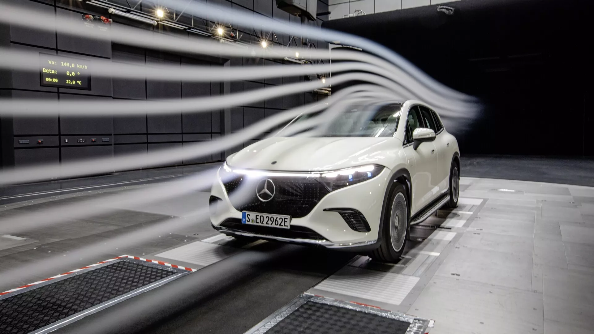 Image d'une Mercedes EQS-SUV 2022 blanche en cours de test dans une installation professionnelle. La voiture, centrée dans le cadre avec un léger angle, montre sa calandre avant, ses phares à LED, son capot, son pare-brise et une partie du côté conducteur. Les lignes de flou de mouvement indiquent des tests aérodynamiques. Design futuriste, grand emblème sur la calandre et environnement de test intérieur.