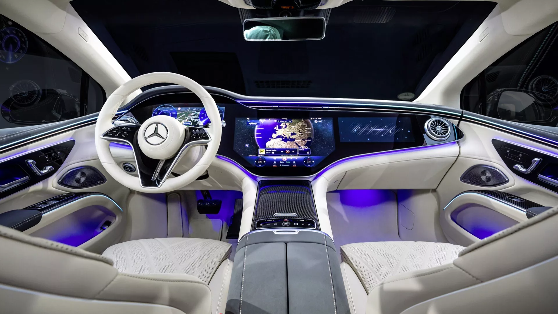 Intérieur du Mercedes-Benz EQS lifting 2024 depuis les sièges arrière soulignant les caractéristiques technologiques et les éléments de luxe, y compris un volant luxueux à deux branches avec contrôles, tableau de bord épuré avec configuration à double écran, console centrale avec commandes sensitives, éclairage d'ambiance bleu, sièges en cuir couleur crème et toit panoramique