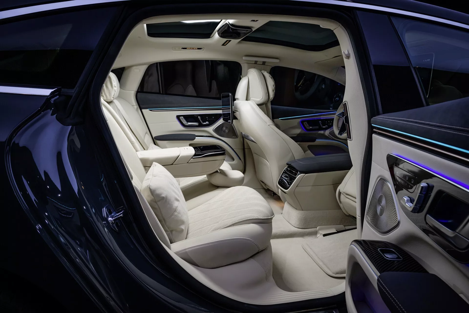 Intérieur du Mercedes-Benz EQS 297 facelift 2024 incluant des sièges en cuir clair sophistiqués, un tableau de bord élégant avec affichage numérique, des panneaux de porte haut de gamme avec commandes, une console centrale étendue, un système de divertissement pour les passagers arrière, un sol en moquette premium et un éclairage d'ambiance dans l'habitacle, vu de l'extérieur avec la porte du passager avant ouverte.