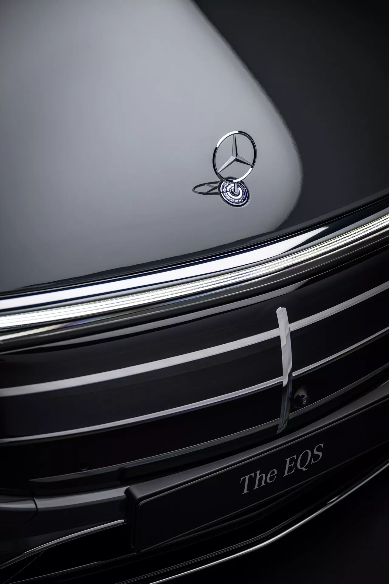 Vue rapprochée de la grille avant et du badge de la Mercedes-Benz EQS 2024, montrant l'emblème chromé contre un capot sombre profilé, avec des lamelles chromées horizontales élégantes et l'inscription 'The EQS' indiquant la conception de véhicules électriques de luxe