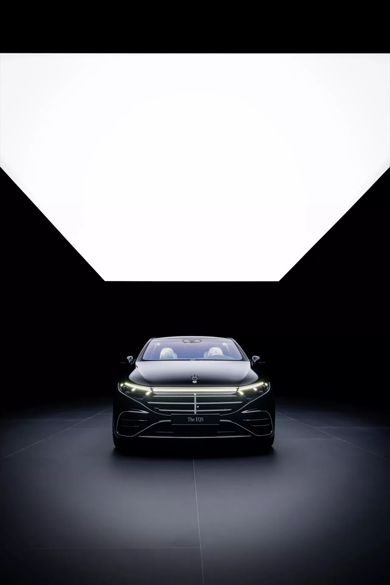 Facelift de la Mercedes-Benz EQS 2024 présentant une vue de face avec des phares LED illuminés et une grille frontale prononcée, capturée sous un angle bas pour mettre en valeur le design élégant et les contours aérodynamiques sur un fond dramatiquement éclairé.