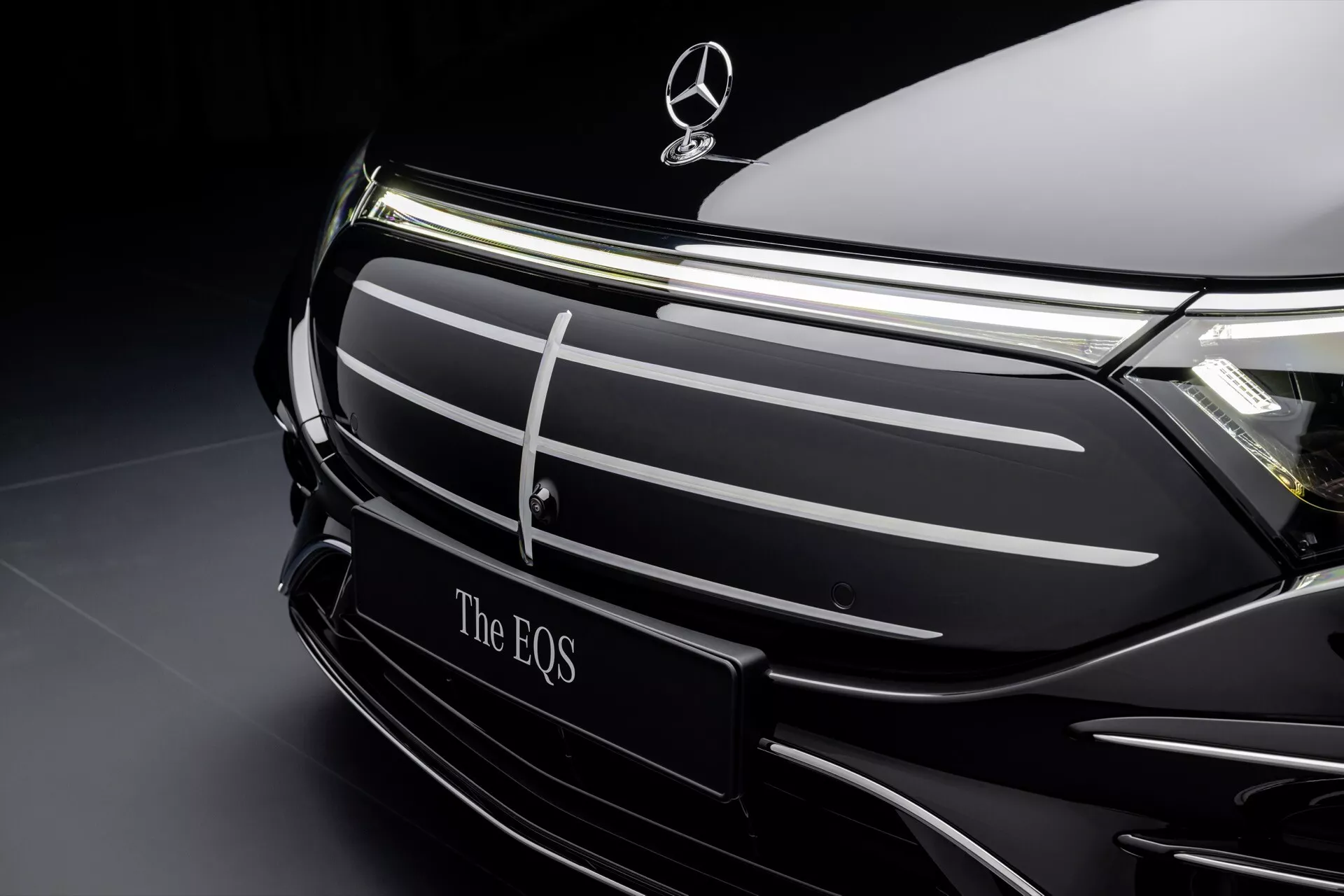 Vue rapprochée de la section avant du modèle Mercedes-Benz EQS 2024 facelift montrant la zone de la grille et l'emblème de la marque sur un fond sombre sophistiqué, mettant en lumière la conception élégante et les caractéristiques de luxe électrique du véhicule