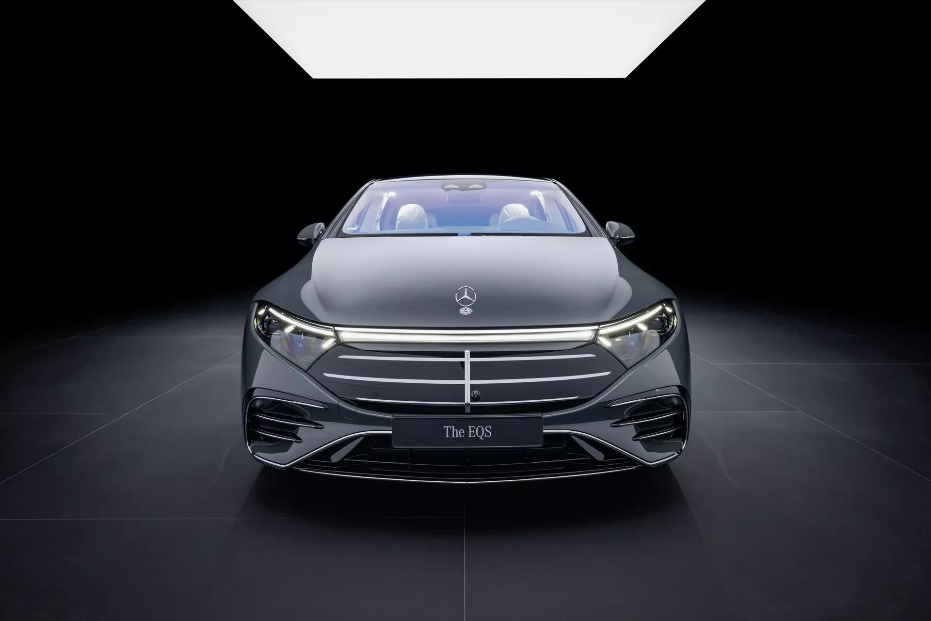 Vue de face de la Mercedes-Benz EQS 2024 (297-Facelift) avec une grille proéminente ornée d'un badge, des phares LED élancés et un design de capot aérodynamique, présentée sous un projecteur dramatique dans un salon auto