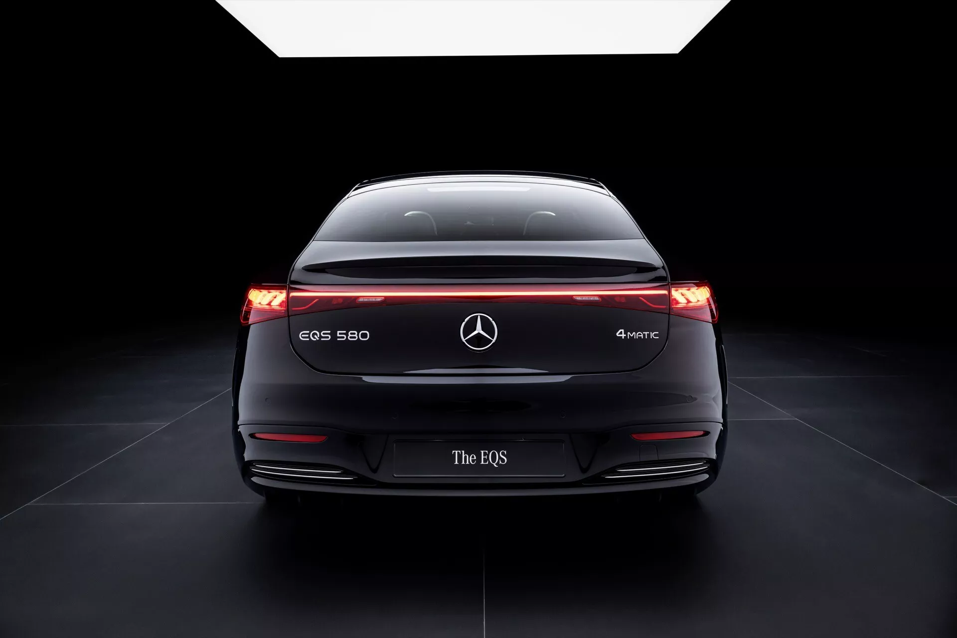 Vue arrière de la Mercedes-Benz EQS 580 4MATIC 2024 mettant en avant sa large stance, feux arrière illuminés avec une bande lumineuse horizontale, désignation du modèle et technologies de sécurité avancées sur un fond dégradé sombre soulignant son aérodynamisme et la conception de véhicule électrique.