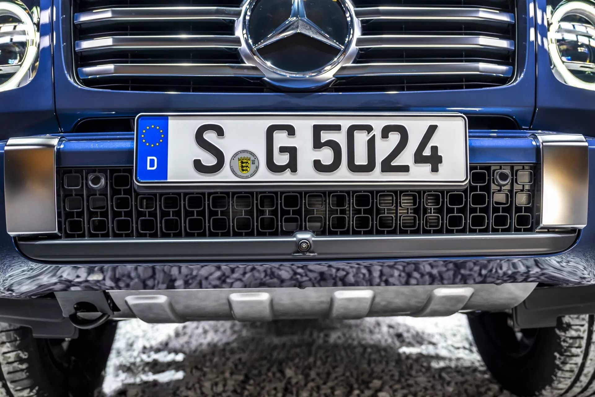 Vue rapprochée du design frontal de la Mercedes-Benz Classe G 2024 montrant la grille avec des rectangles noirs encadrés de chrome, plaque d'immatriculation européenne, pare-chocs avant robuste avec plaque de protection métallique en bas, et l'insigne central Mercedes-Benz pouvant intégrer une technologie de capteurs, peint en bleu foncé, indiquant un SUV de luxe ou haute performance en état immaculé.