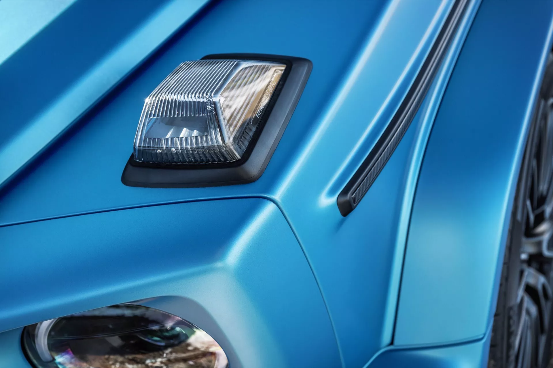 Gros plan sur des détails du Mercedes-Benz G-Klasse 2024 comprenant une prise d'air sur le capot, des lignes de carrosserie anguleuses, une peinture bleue métallisée et une aération latérale de l'aile, mettant en évidence les éléments de design et de technologie